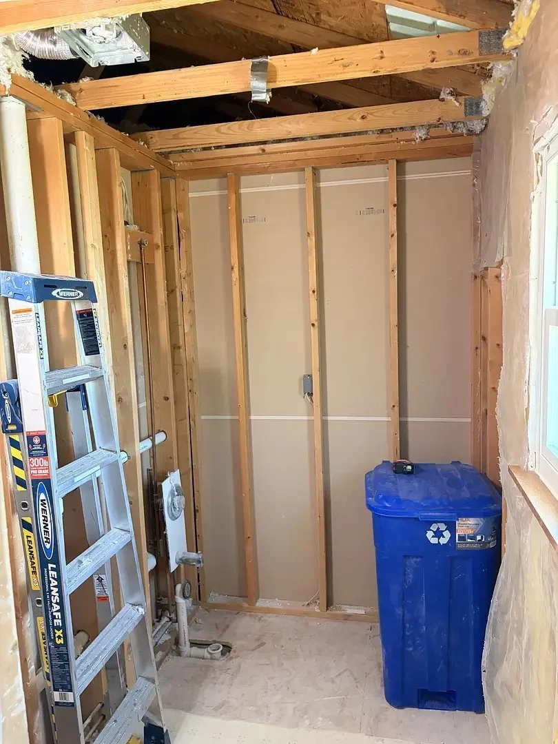 Unfinished room with exposed wooden studs, a ladder, pipes, and a blue recycling bin. Ceiling partially open