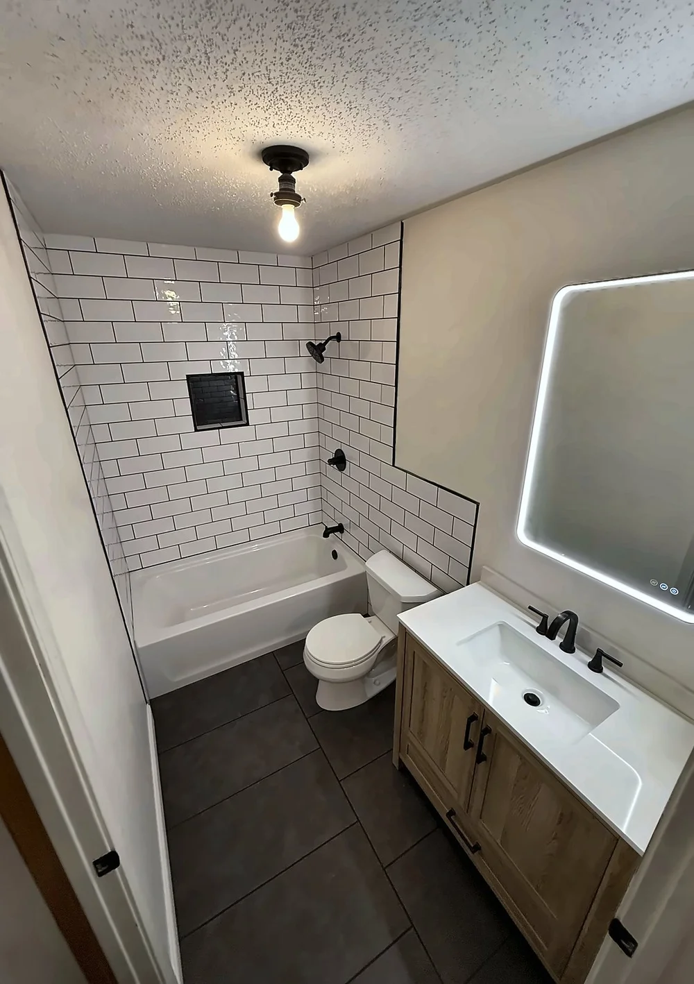 Remodeled hall bathroom with white subway tile, a tile niche, matte-black fixtures, a wood vanity and a backlit LED mirror
