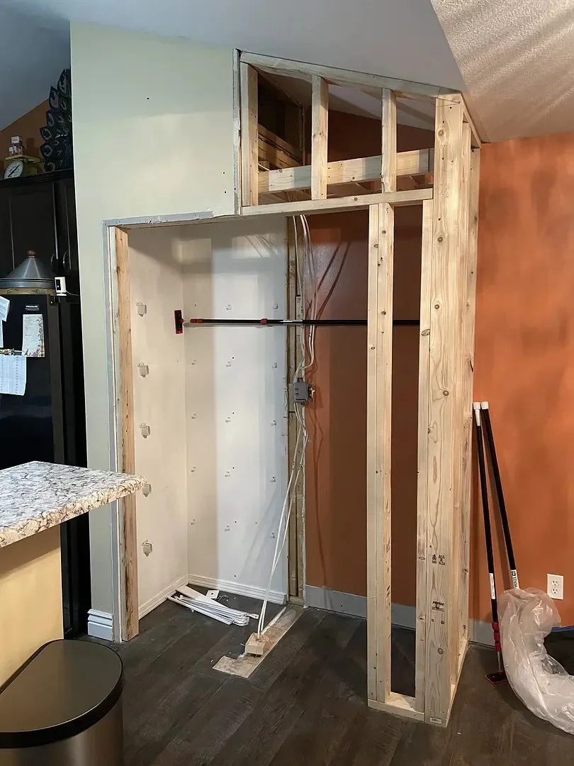 Framed wooden structure in a kitchen under construction, beside a countertop and fridge. Wiring is visible