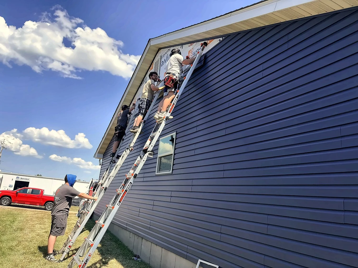 HP Pros crew on ladders hanging Midnight Blue vinyl siding on the side of a ranch home, with a crew member on the ground in a cooling towel
