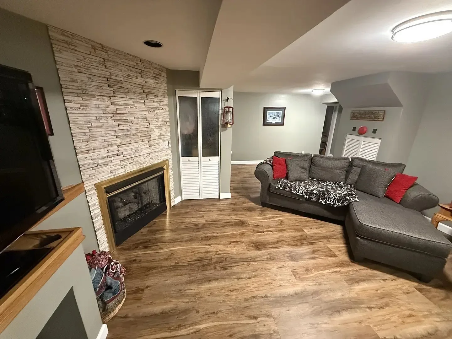 A cozy living room with a stone fireplace, gray sectional sofa, red cushions, and wooden floor