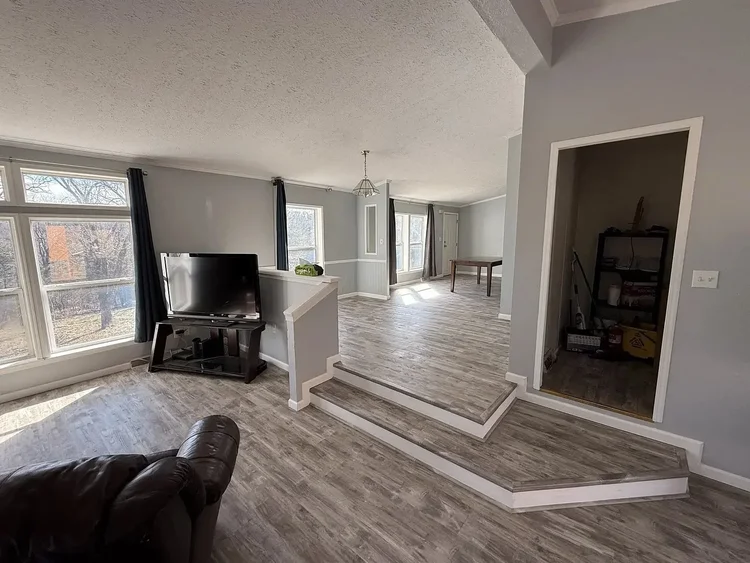 Spacious living room with TV, steps, large windows, and light gray walls. Sunlit area with wooden floors