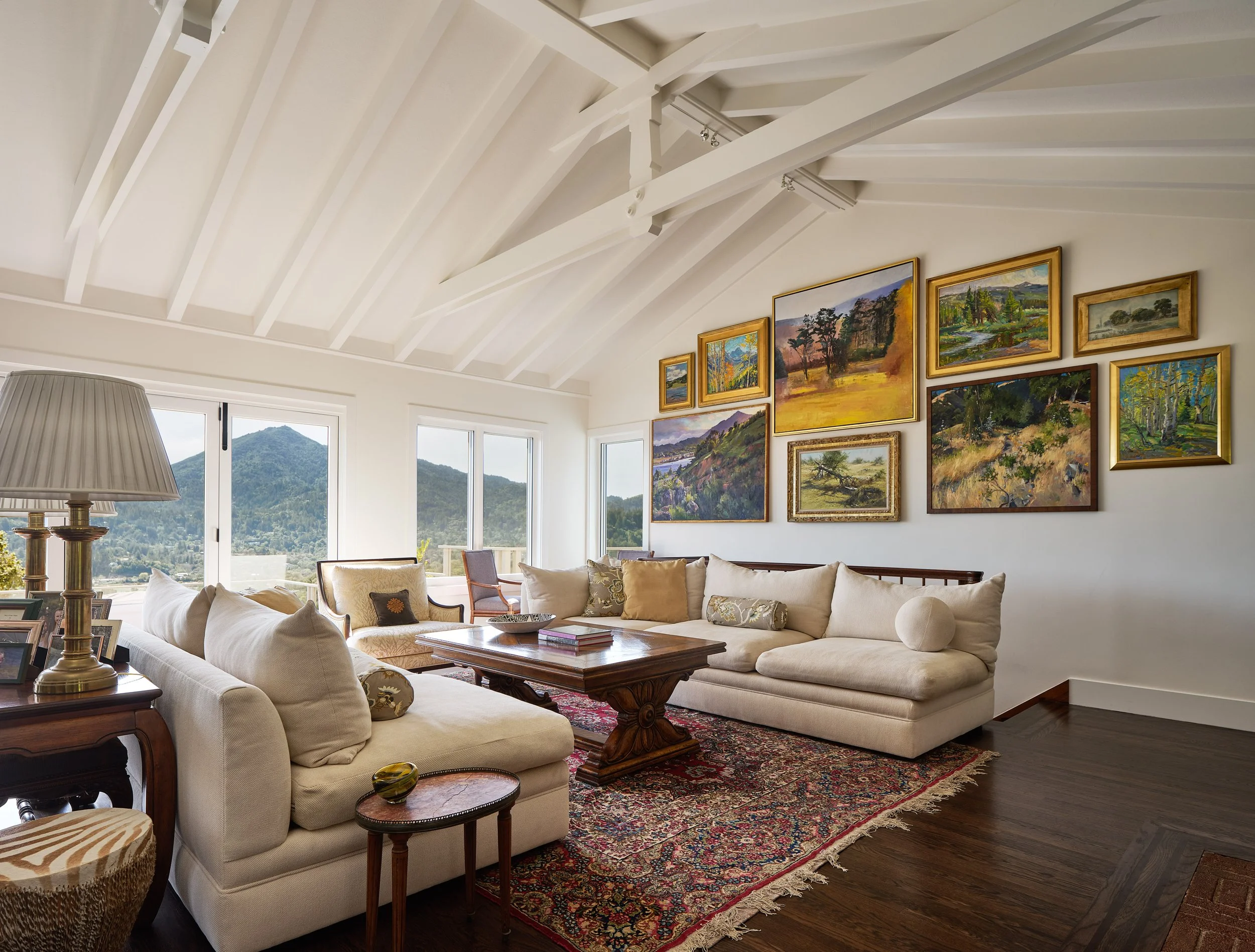 Living room with beige sofa, wooden coffee table, colorful area rug, wall art, large windows showing mountain view, and a lamp.