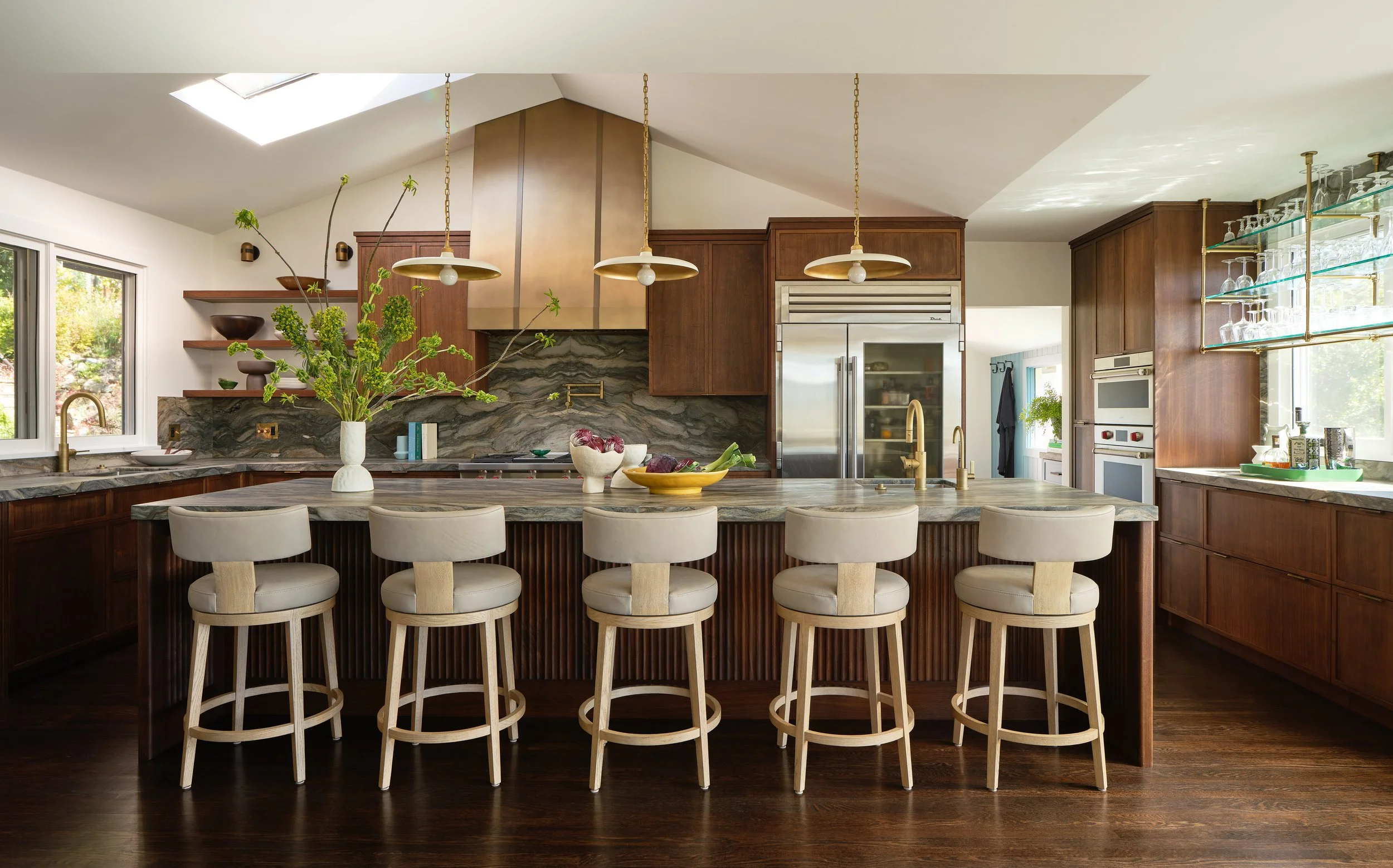 Modern kitchen with a large marble island, wood cabinets, and gold fixtures, decorated with a tall vase of green branches and various bowls of food.