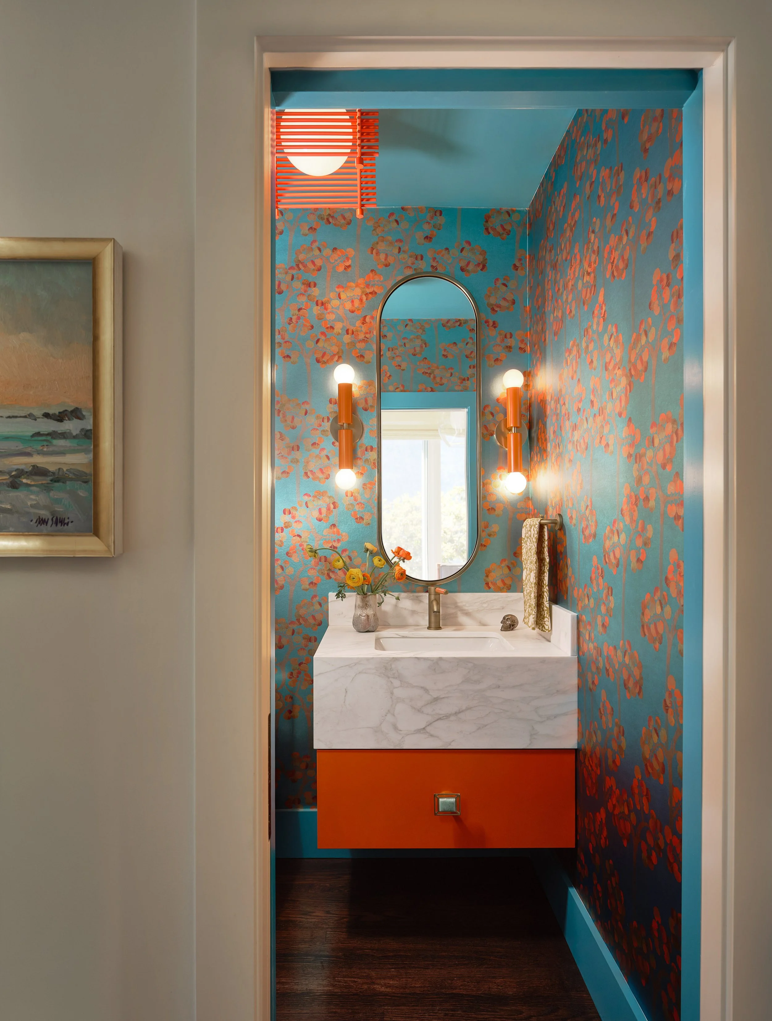 A photograph of a small bathroom with bold, colorful wallpaper featuring orange trees on a blue background. The bathroom has a white marble sink with a gold faucet, flanked by two modern wall sconces with white globes and orange holders. There is a l