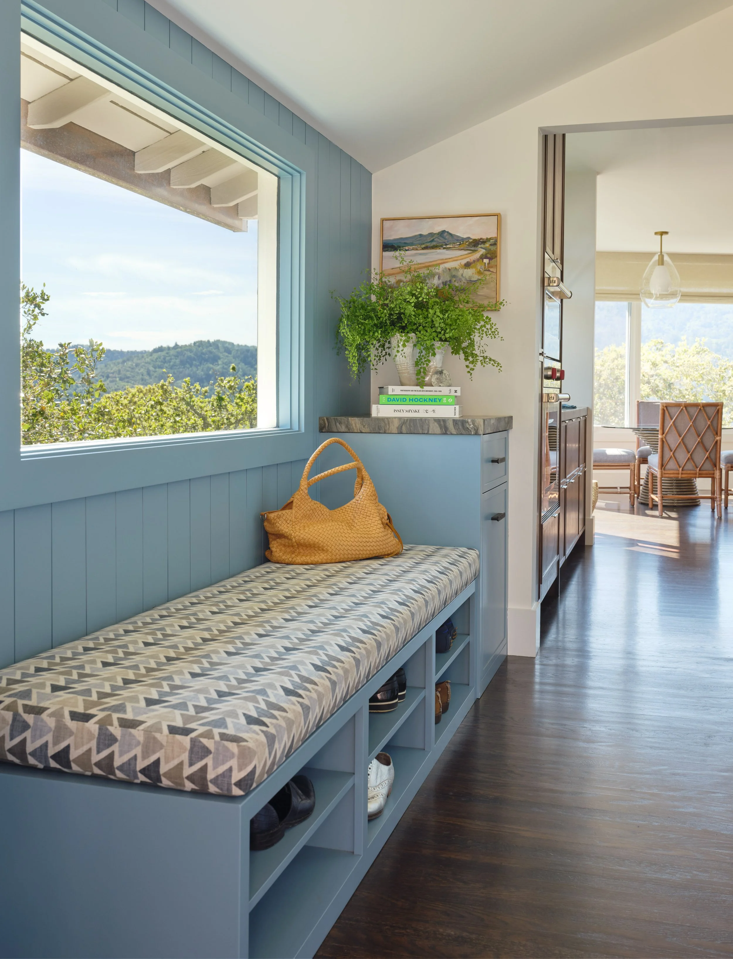 A built-in bench with a geometric-patterned cushion, a yellow handbag on top, and open cubbies beneath filled with shoes, in a sunlit room with a large window and a view of trees and hills.