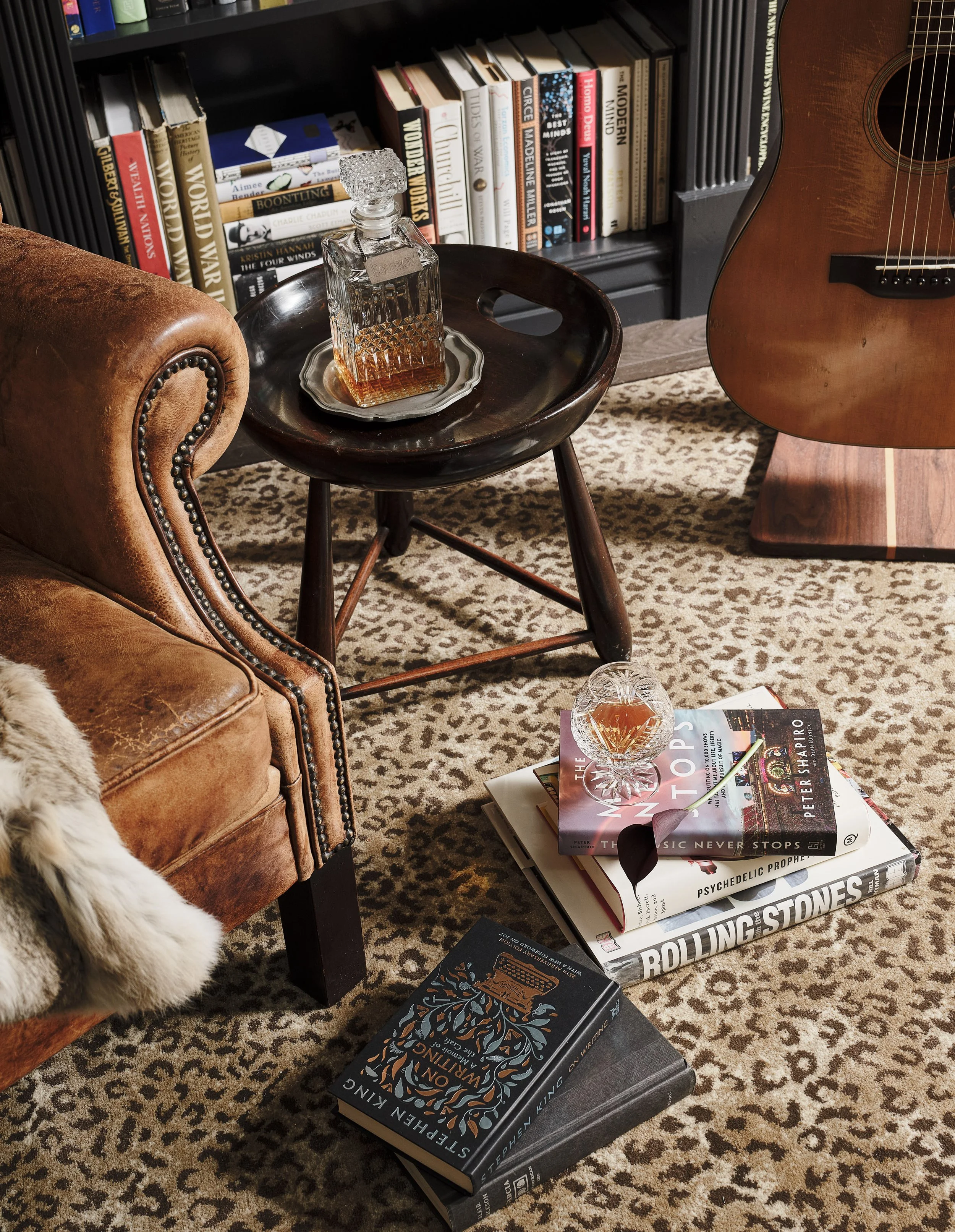 A cozy reading corner with a well-stocked bookshelf, a wooden side table with a glass decanter of whiskey on a tray, a brown leather armchair with nailhead trim, a guitar leaning against the wall, a small side table with a decorative glass bowl and m