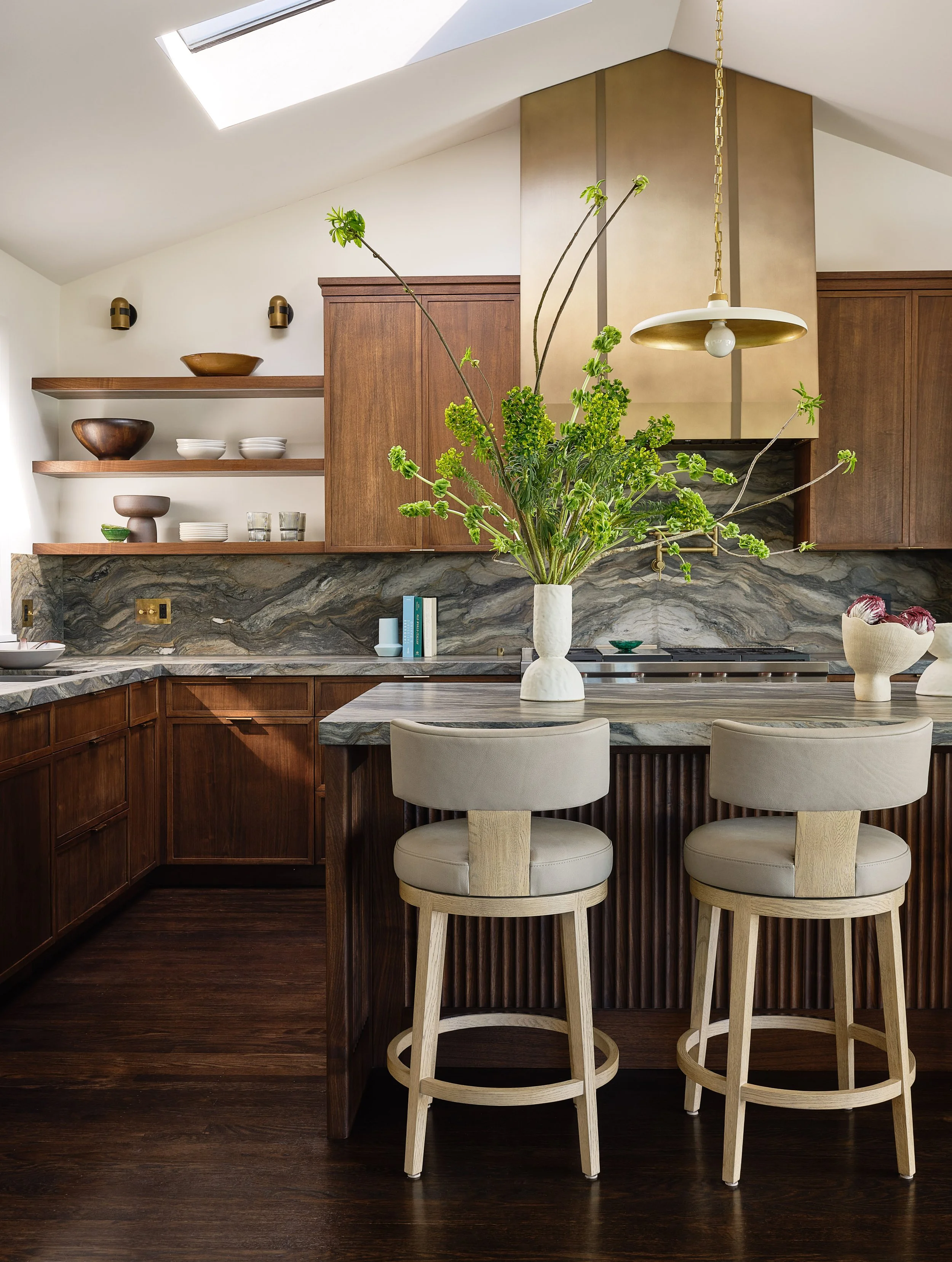 A modern kitchen with wooden cabinets, open shelving with bowls and dishes, a marble countertop, and two white bar stools with wooden legs at a marble-topped island. A large vase with green flowers is on the island, and overhead pendant lighting is v