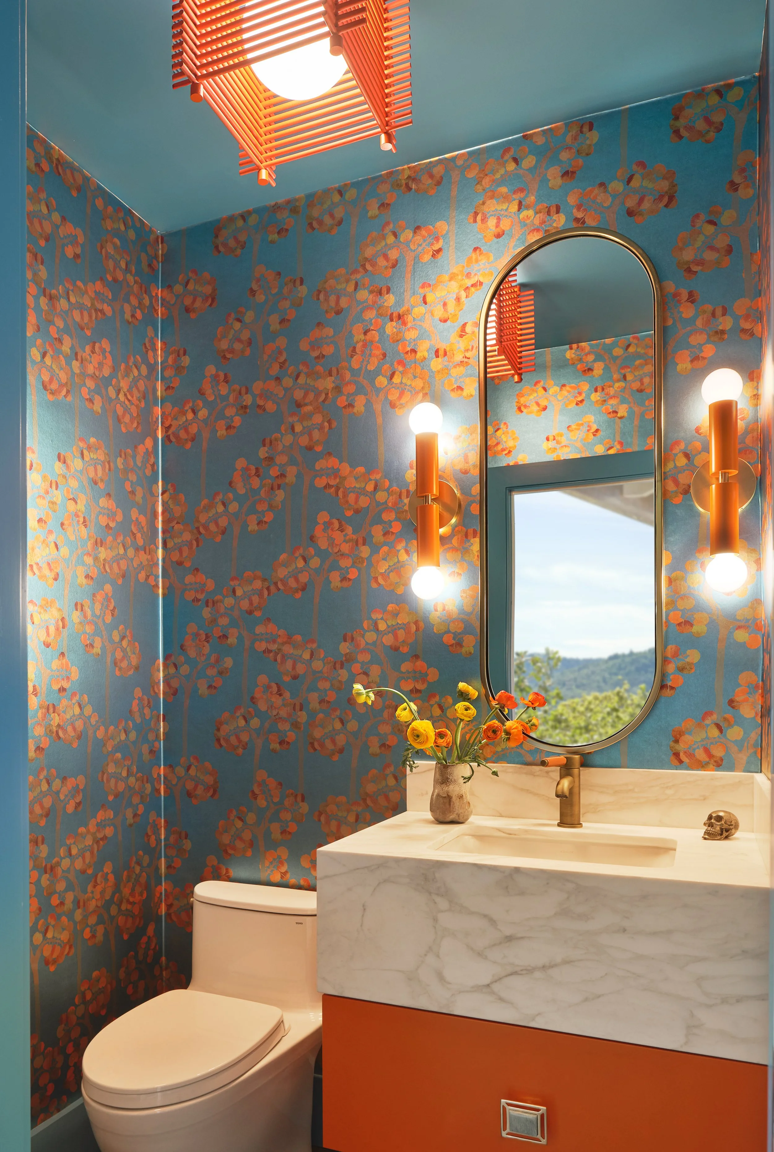 A small bathroom with blue floral wallpaper, a round mirror, a beige marble sink with a brass faucet, a vase with yellow and orange flowers, a lighting fixture with two bulbs and orange accents, a window showing a scenic outdoor view, a skull ornamen