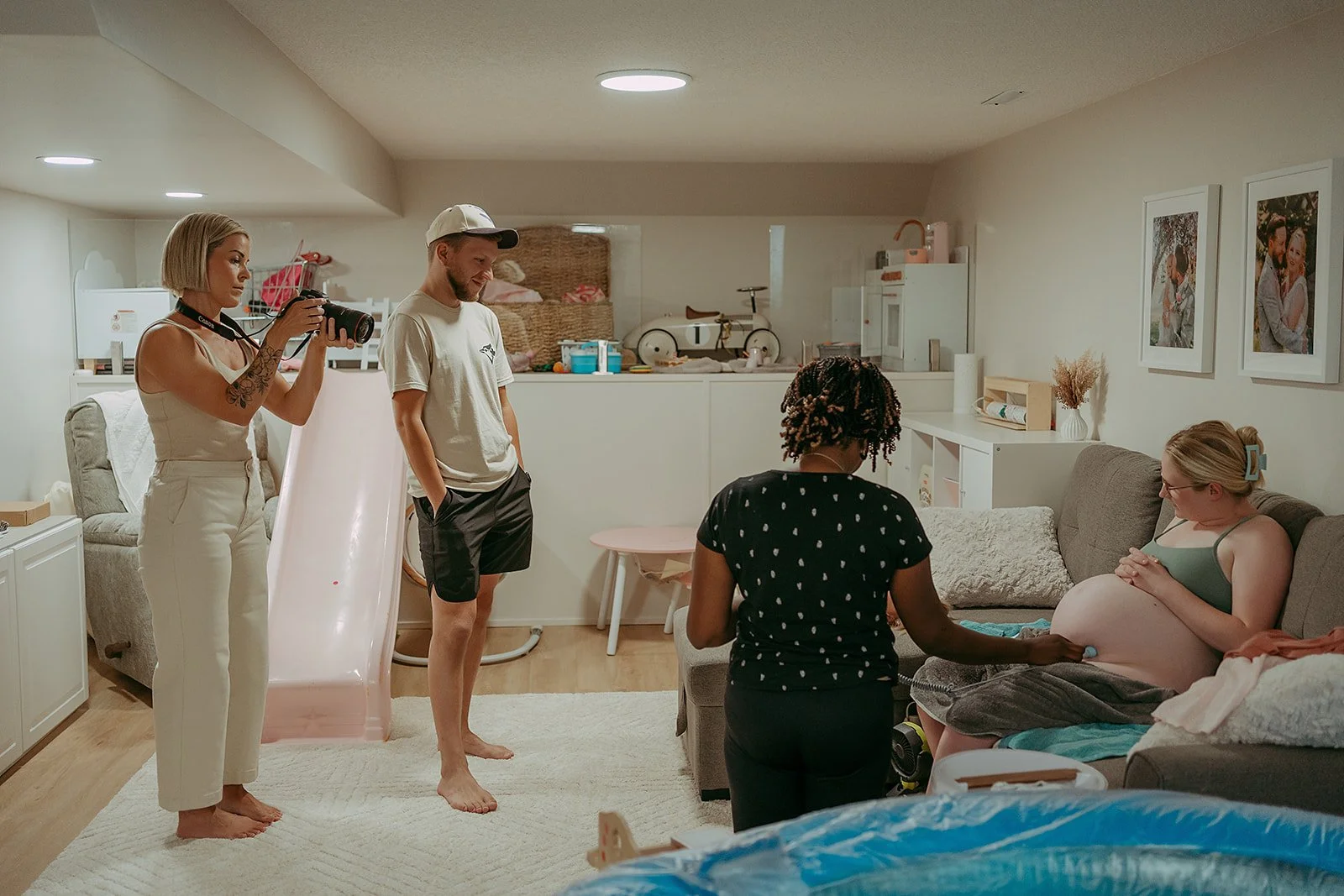 A woman is pregnant and sitting on a sofa in a living room while a midwife checks her belly, with two people taking photos in the background.