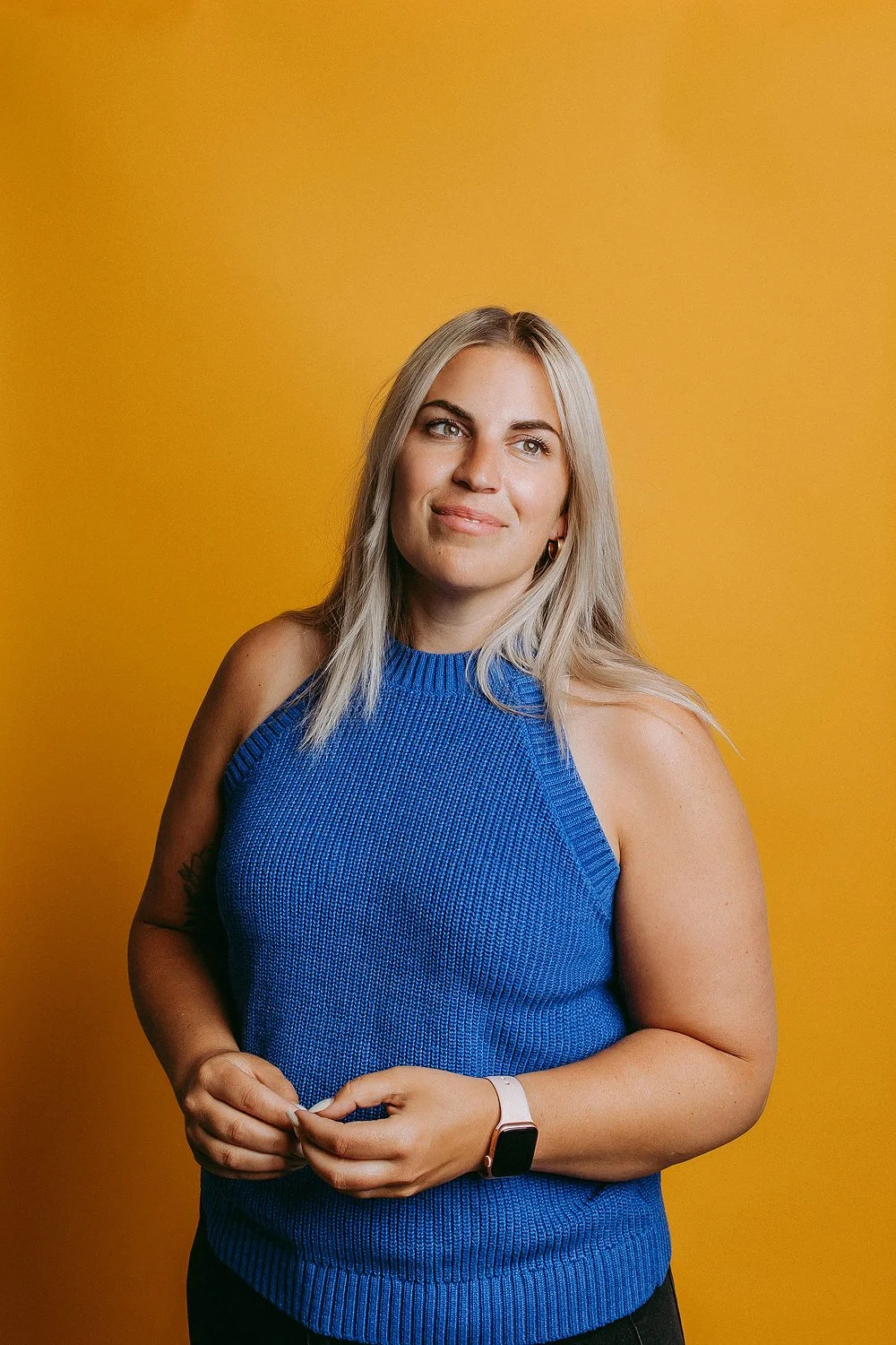 A woman with blonde hair in a royal blue sleeveless sweater stands against a yellow backdrop, with a smartwatch on her left wrist, looking slightly upwards with a slight smile.