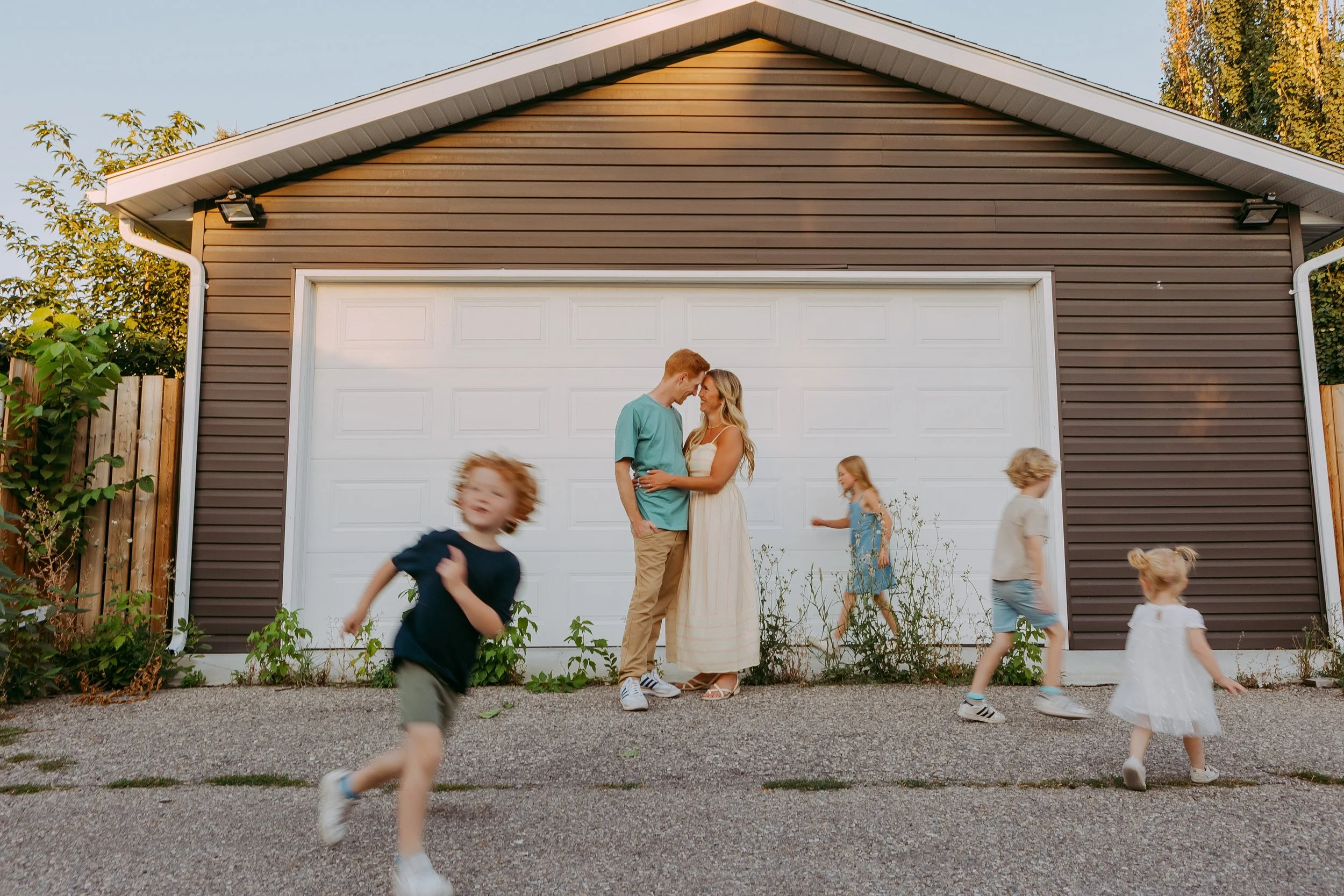 A young couple stands close together, touching foreheads and smiling, with four children playing and running in front of a white garage door on a sunny day.