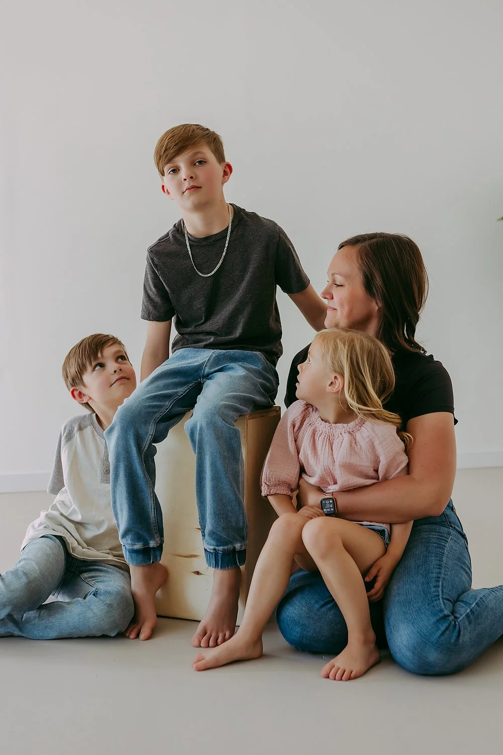 A woman with three children, one boy sitting on her lap, another boy sitting on the floor, and a girl sitting on her lap, in a minimalist room with white walls.