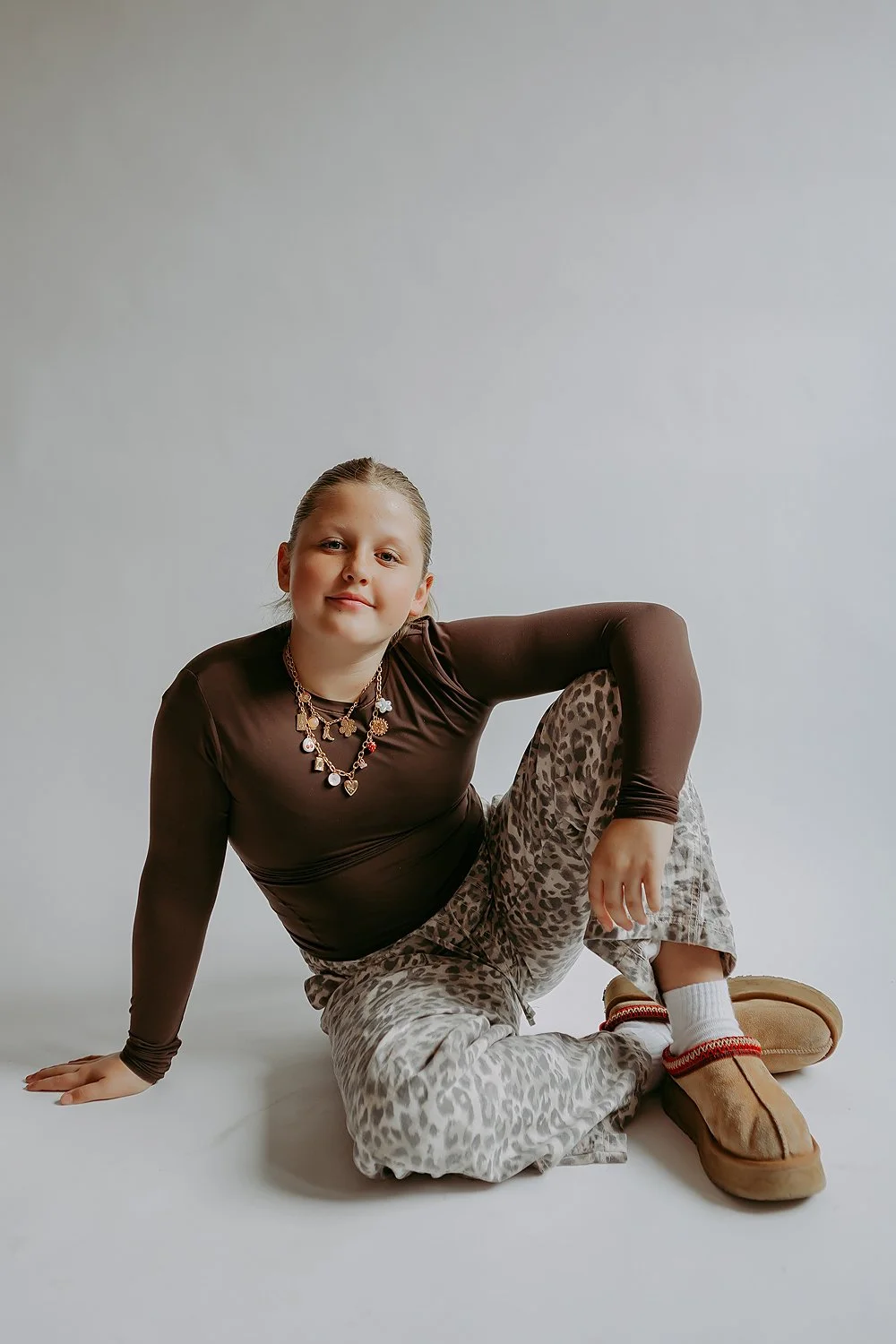 A young girl with blonde hair pulled back, wearing a brown long-sleeve shirt, leopard-print pants, beige slippers, and layered necklaces, sitting on the floor against a plain white background.