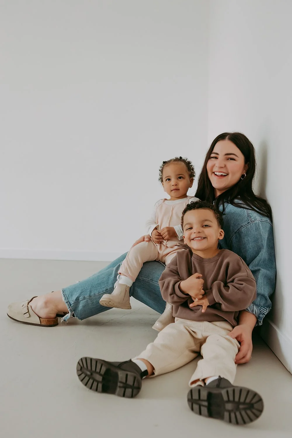 Smiling young woman with two children sitting on the floor against a white wall.