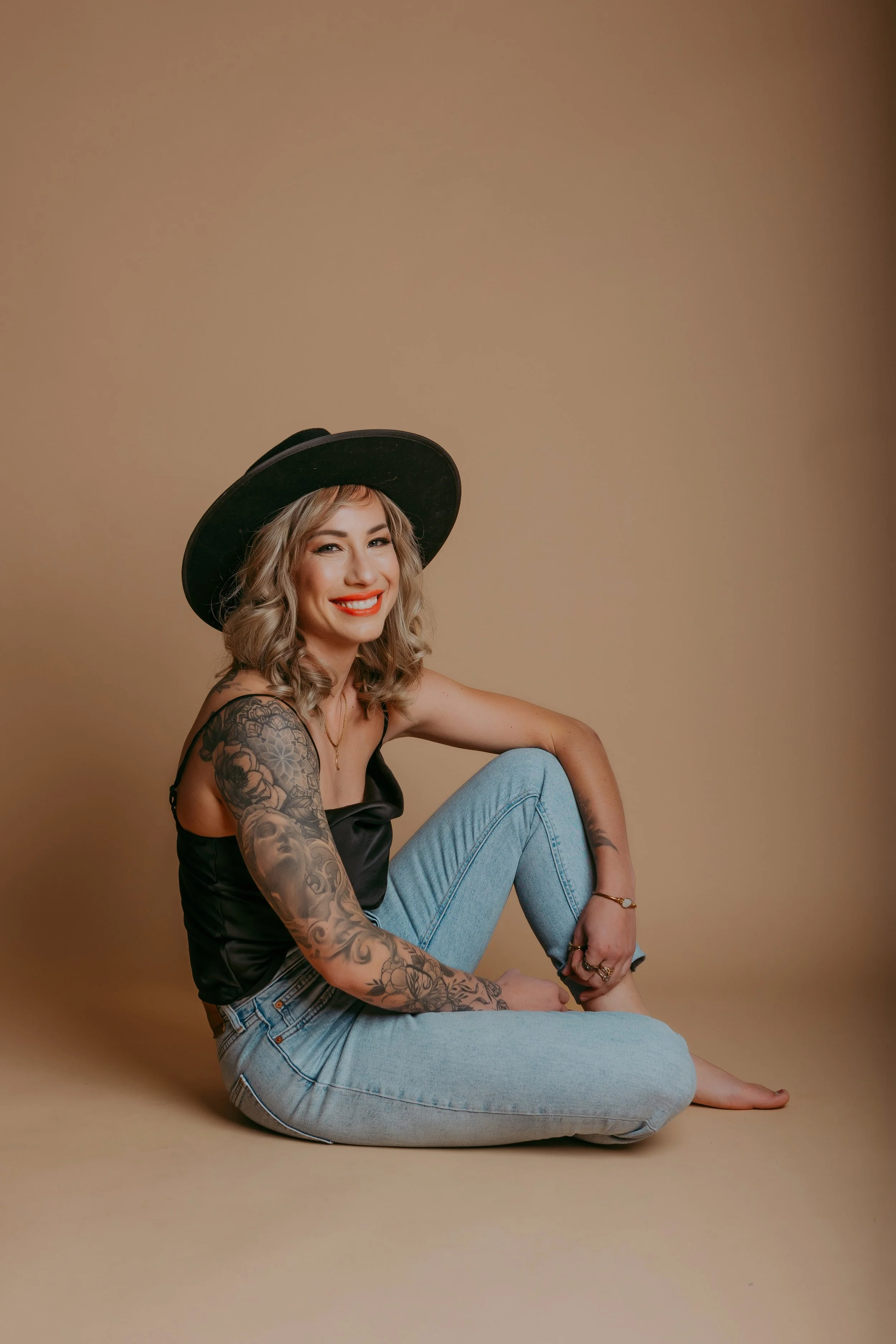 A smiling woman with blonde hair, tattoos on her arms, wearing a black wide-brimmed hat, black silk tank top, and light blue jeans, sitting on a beige background.