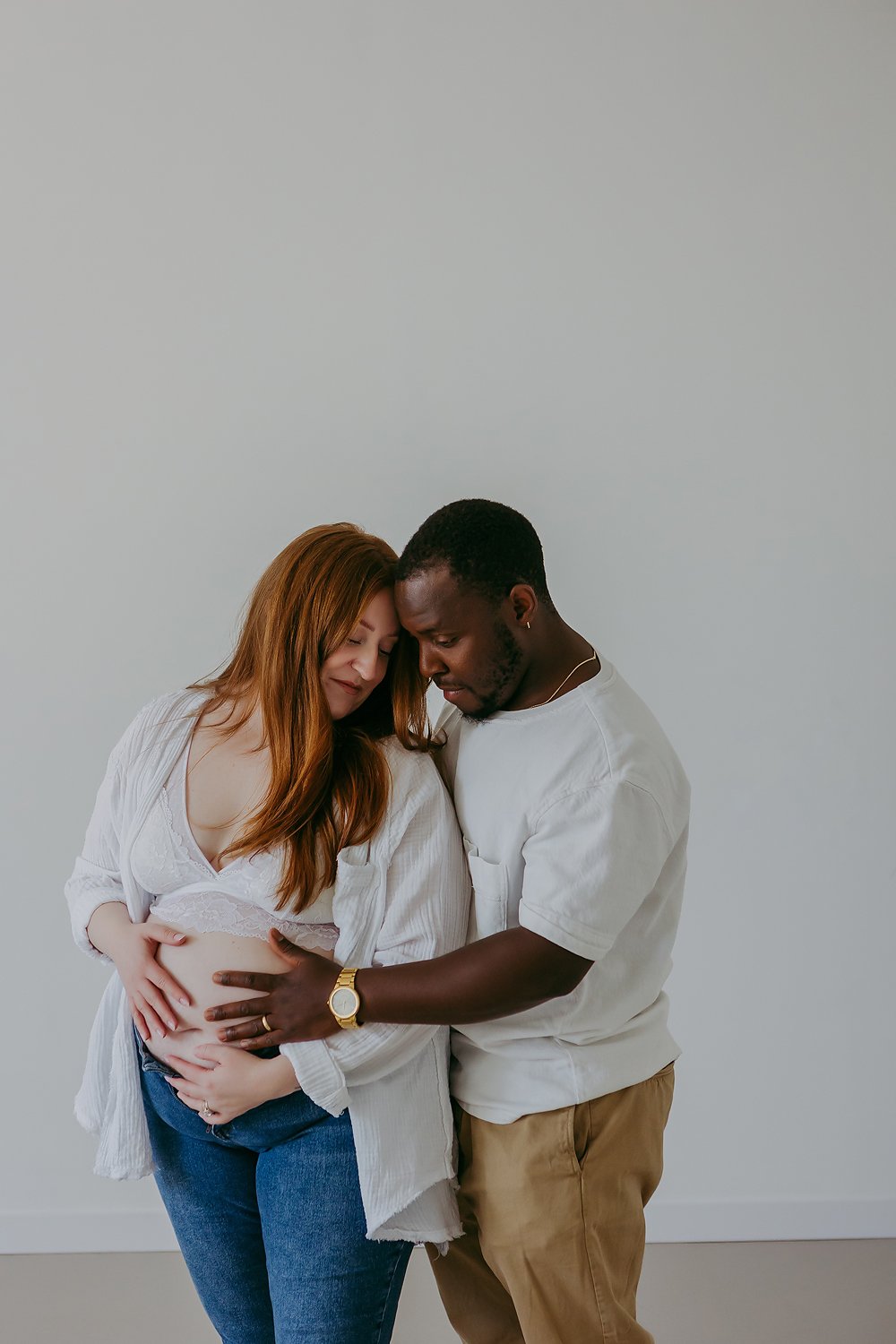 Pregnant woman with red hair and their partner gently touching her belly, both with eyes closed and leaning their foreheads together, standing against a plain light gray background.