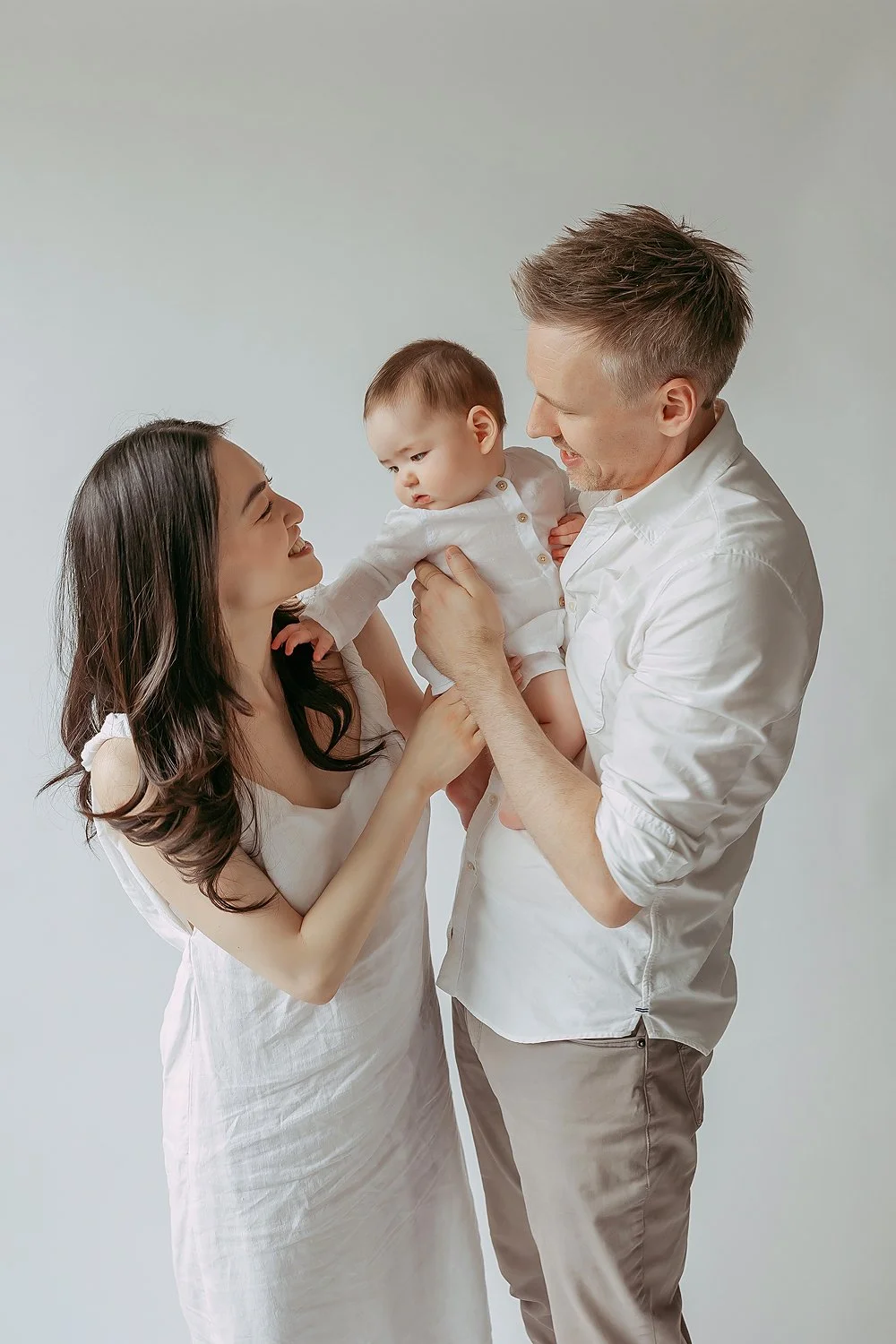 A joyful family of three with a woman, a man, and a baby, smiles and looks at each other against a plain light background.