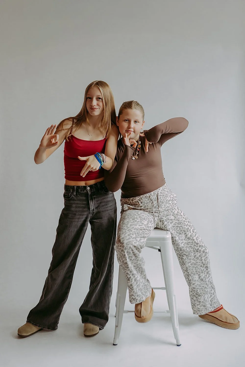 Two teenage girls posing playfully in front of a plain background. One standing with a peace sign and the other sitting on a stool, making a gesture with her finger near her mouth.