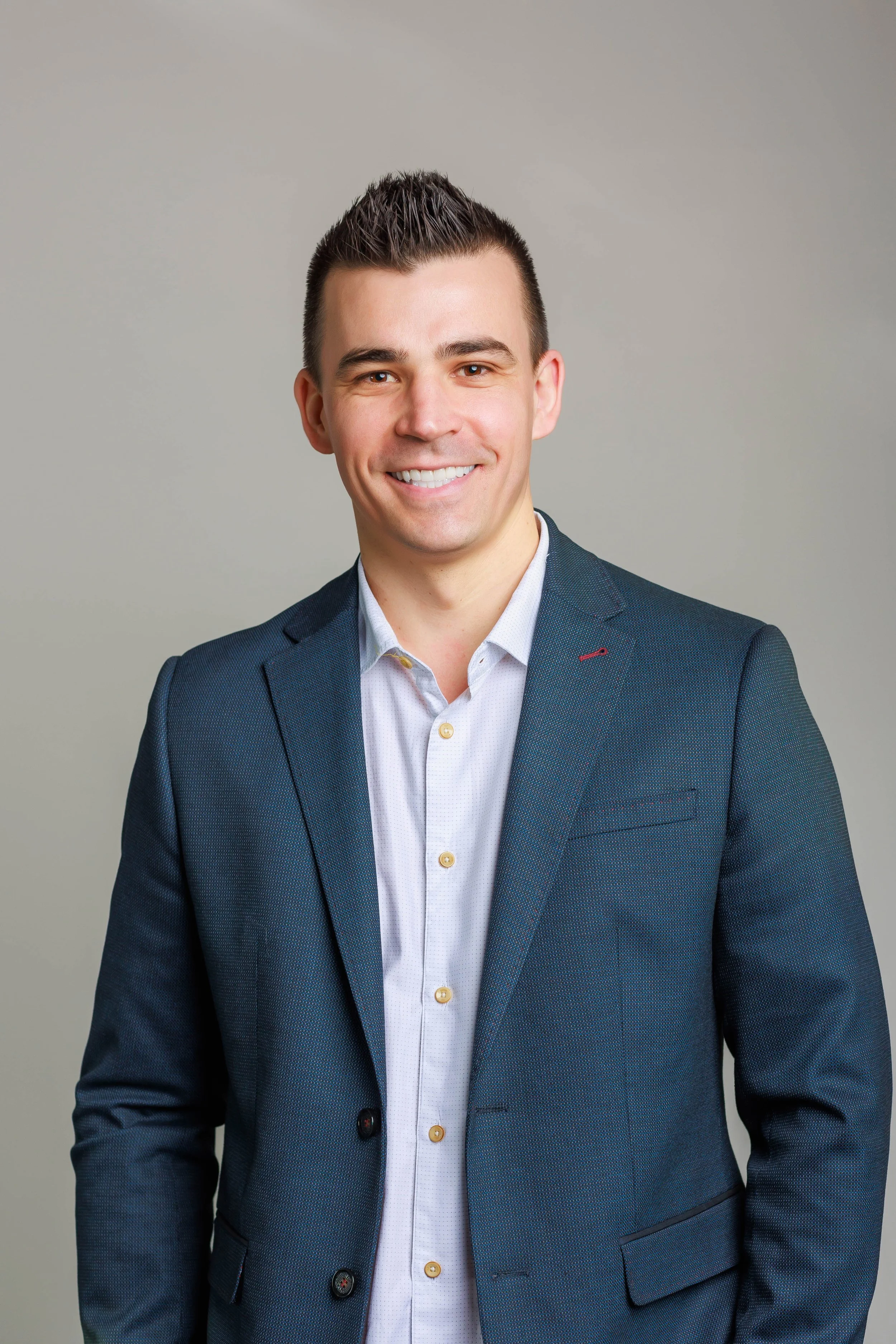 Portrait of a young man wearing a dark suit and white shirt, smiling against a plain grey background.