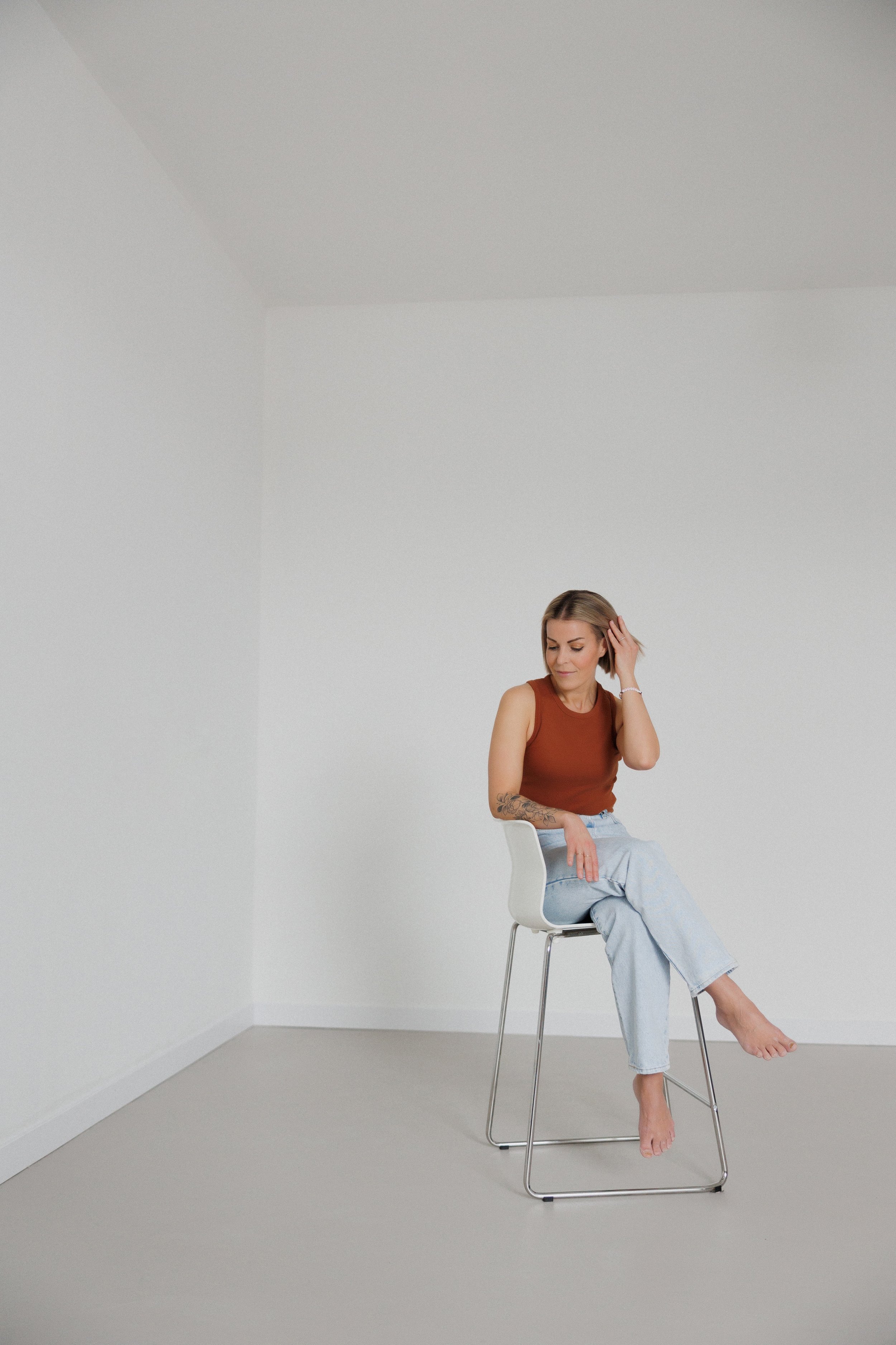 A woman sitting on a white chair in a plain, white room, looking down and touching her hair.