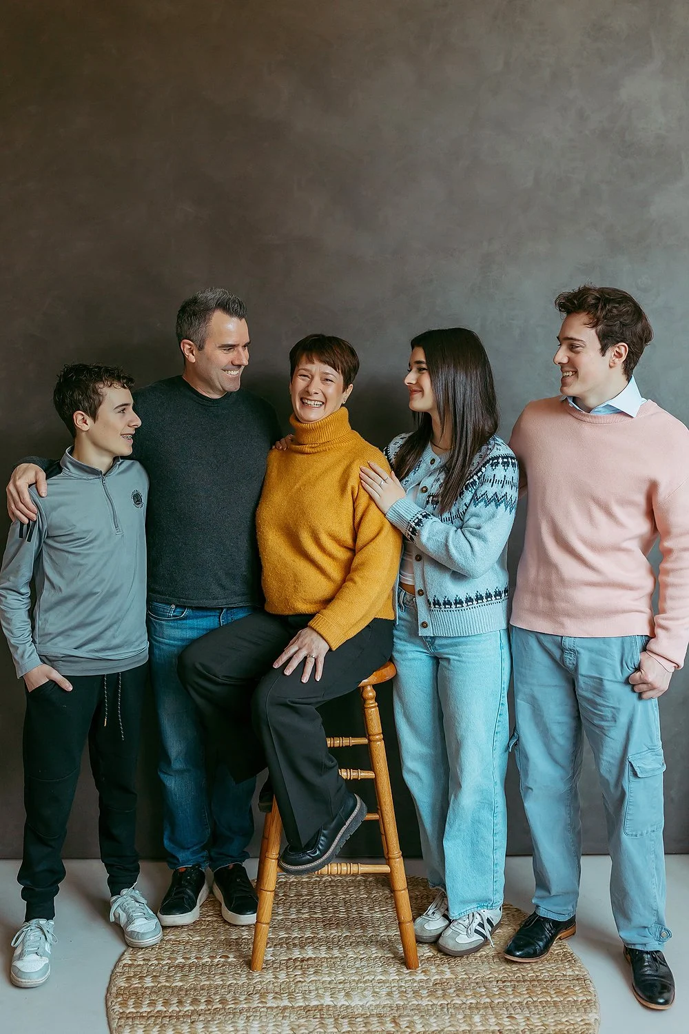 Family of five happily smiling and interacting, with one woman sitting on a wooden stool, others standing around her against a gray wall.