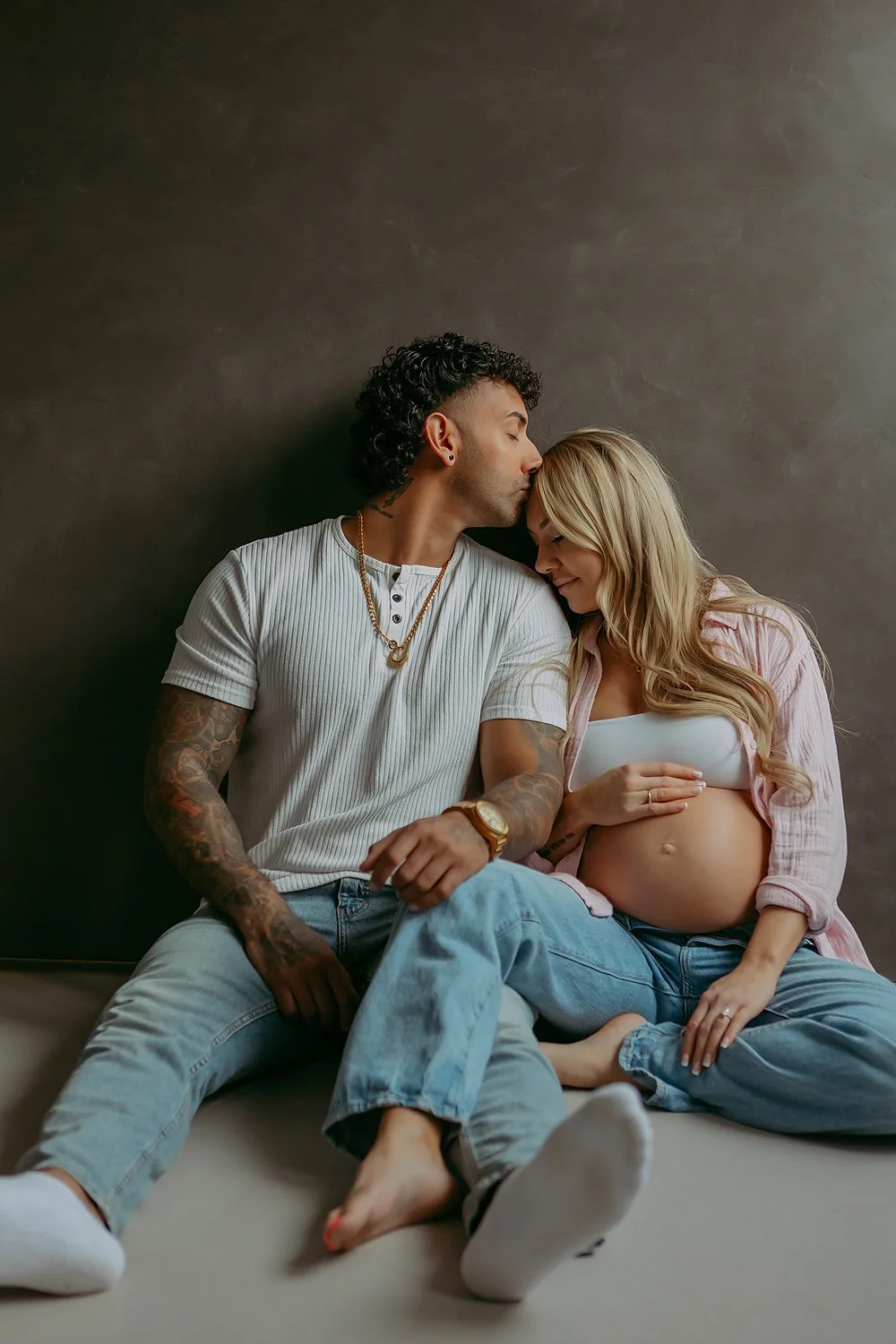 A couple sitting on a bed, with the woman showing her pregnant belly, as they touch foreheads and smile.