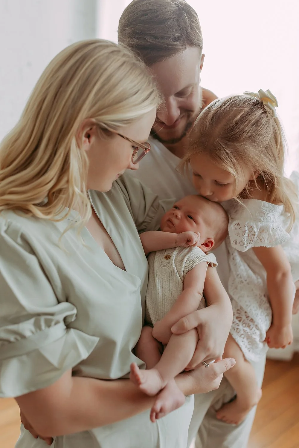 A family of four, including two young children, a baby, and both parents, sharing a loving moment in a studio, with everyone focused on the baby.
