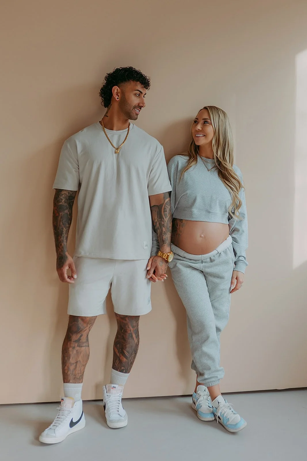 A man and woman holding hands and smiling at each other, standing against a plain beige wall. The man is dressed in casual light-colored clothing with tattoos on his arms and legs, while the woman has long hair, an exposed pregnant belly, and is wearing a gray sweatshirt and sweatpants.