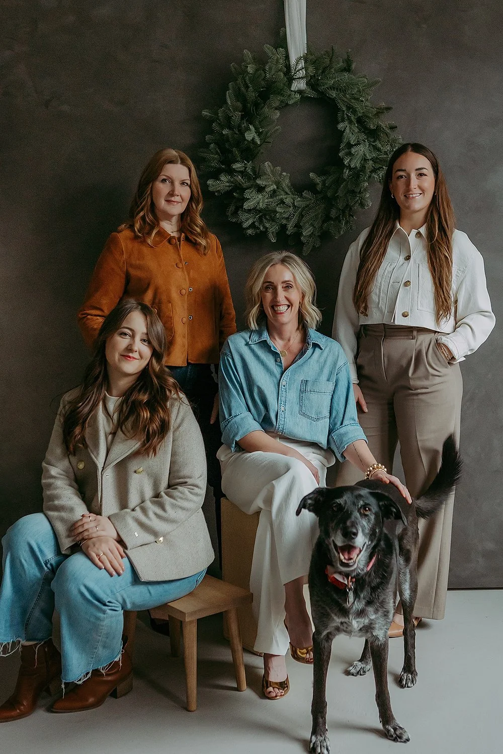 Five women and a dog posing in a studio with a black wall and wreath decoration in the background.