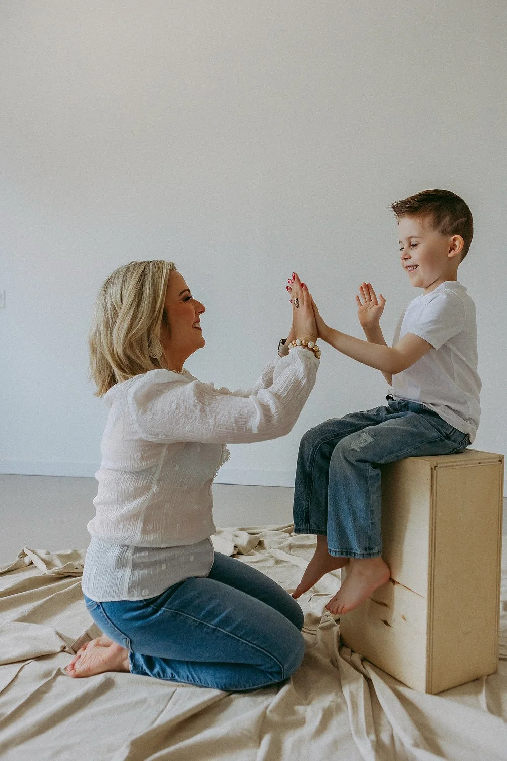 A woman and a young boy are sitting on a beige sheet in front of a plain white wall, giving each other high fives and smiling. The woman is kneeling on the sheet, wearing a white blouse and blue jeans. The boy is sitting on a wooden box, wearing a wh