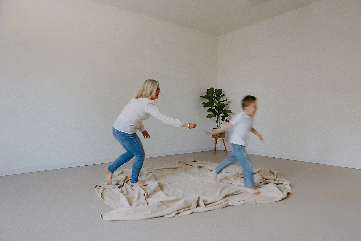 Woman and young boy playing on a bed covered with a beige sheet in an empty room with white walls and a potted plant.