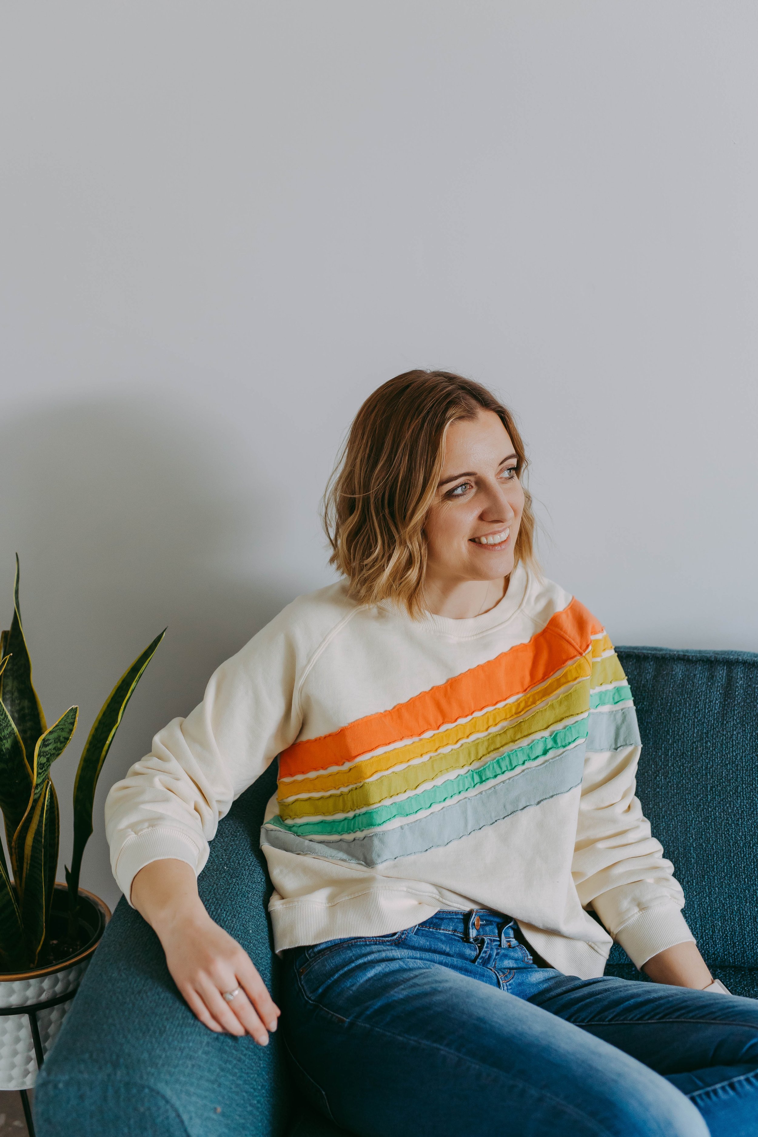 A woman with shoulder-length wavy hair sitting on a blue couch, smiling, wearing a cream sweatshirt with colorful rainbow stripes across the chest, in a room with a white wall and a potted plant beside her.