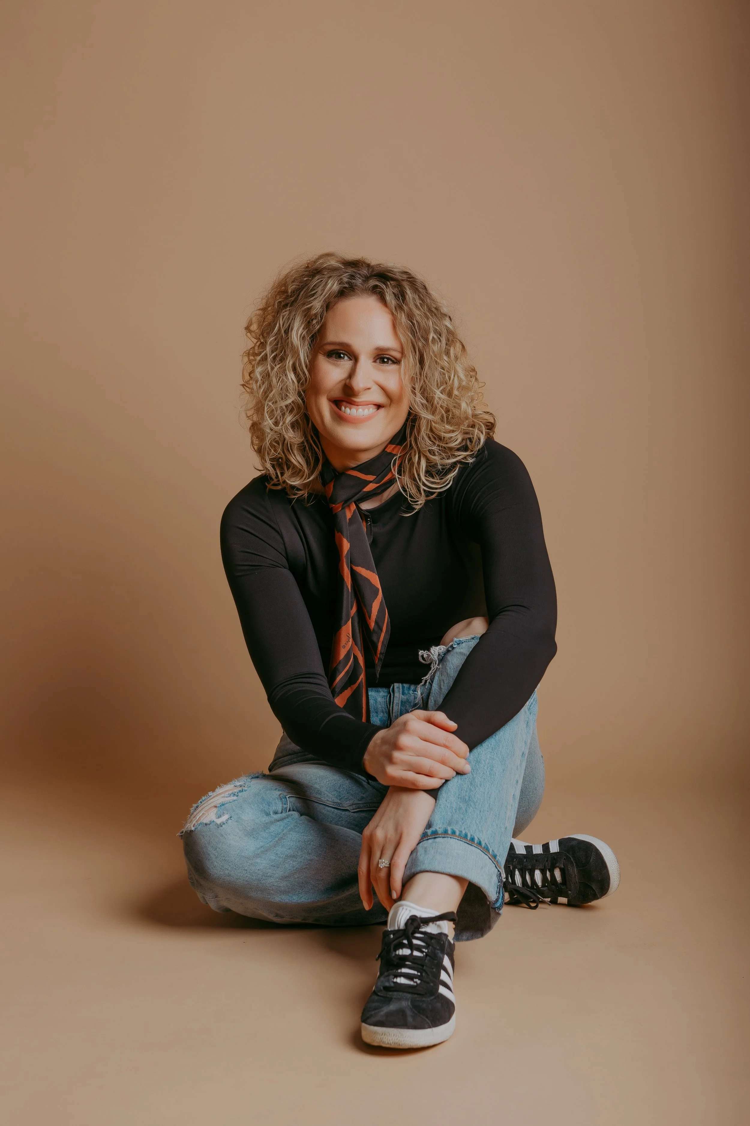A woman with curly blonde hair sitting on the floor against a beige background, smiling at the camera. She is wearing a black long sleeve shirt, a colorful scarf, ripped jeans, and black sneakers with white stripes.