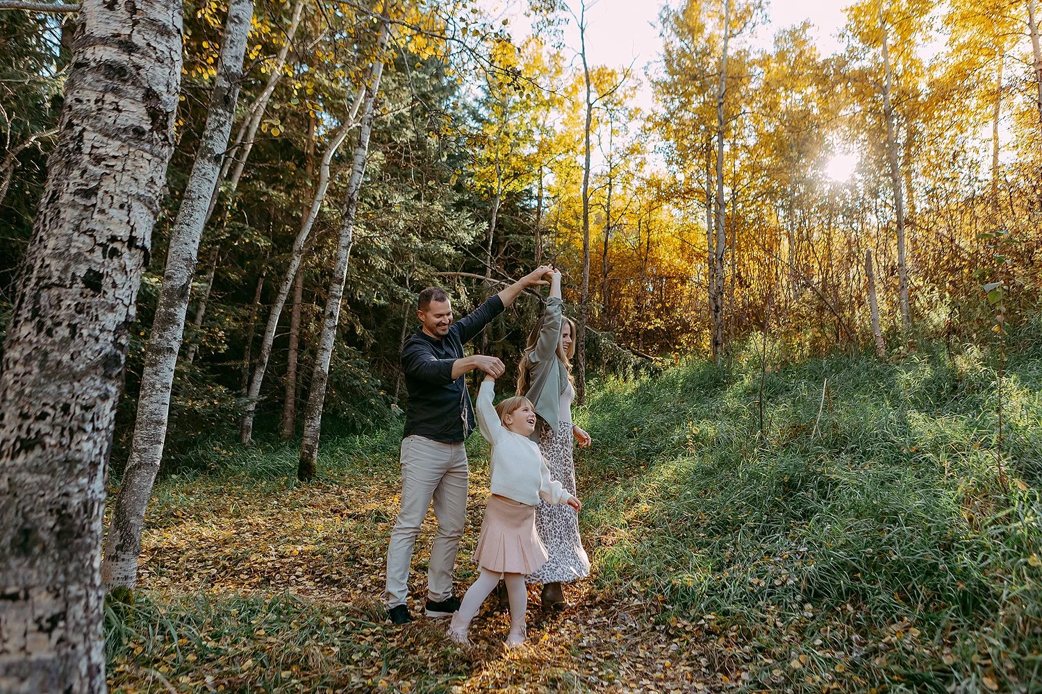 Family of three enjoying a walk in the woods during fall, holding hands and smiling, with sunlight filtering through trees with yellow leaves.