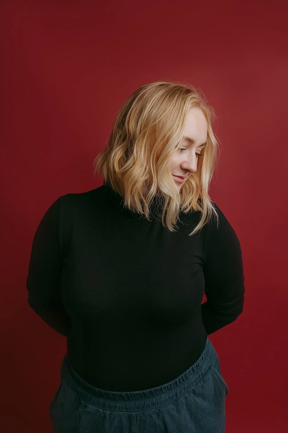 A woman with shoulder-length wavy blonde hair, smiling with eyes closed, wearing a black turtleneck and dark pants or shorts, standing against a solid red background.