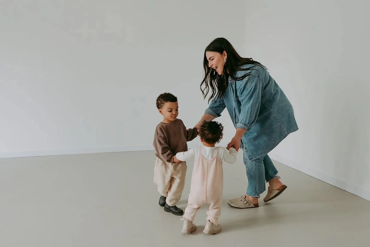 A woman in a denim jacket and jeans playing with two young children in a spacious, empty room.