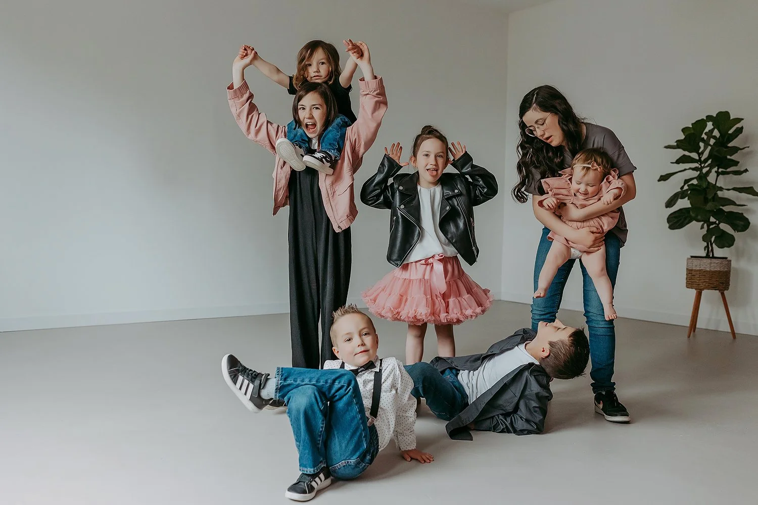Six children with a woman posing playfully in a minimal room with a potted plant in the background.