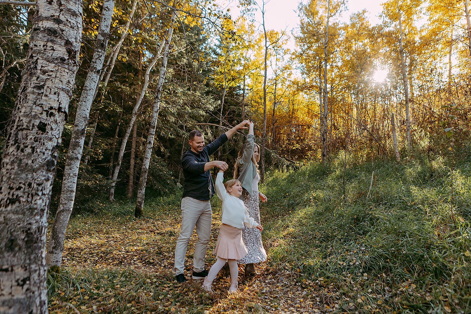 A family of three, a man, a woman, and a girl, walking through a wooded forest during autumn, with sunlight shining through the trees.