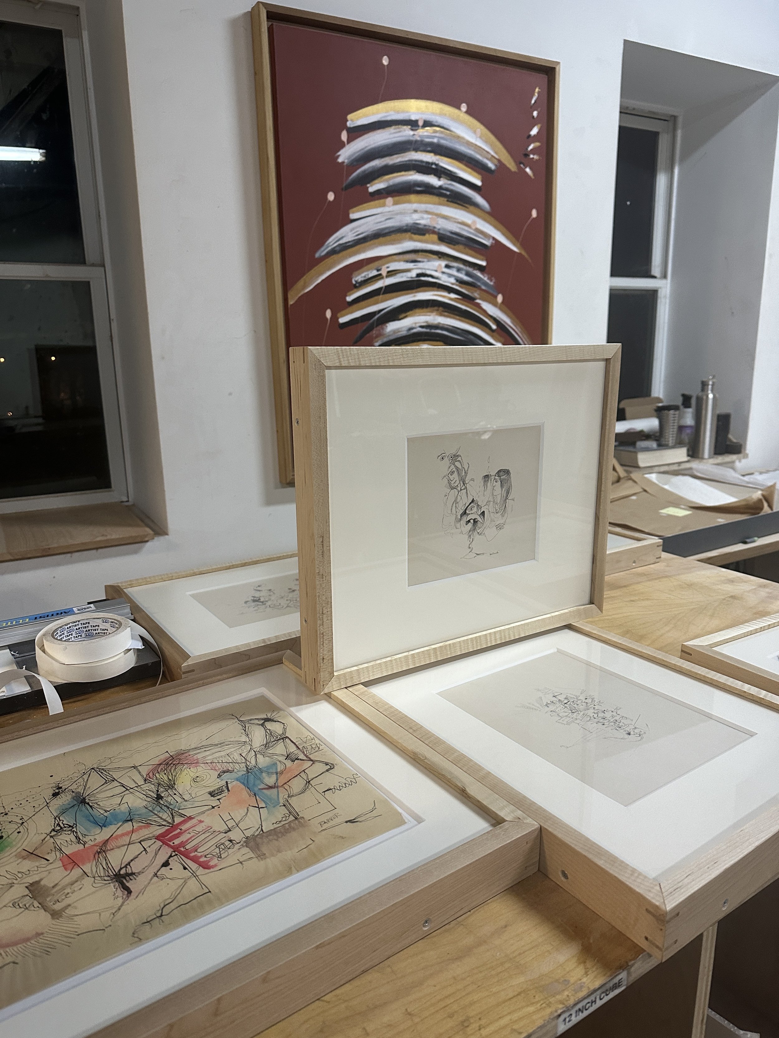An art studio workspace showcasing a large abstract painting with curved brushstrokes in black, white, and gold on a reddish background, and various smaller framed drawings and sketches on a wooden table, with tools and supplies visible in the backgr