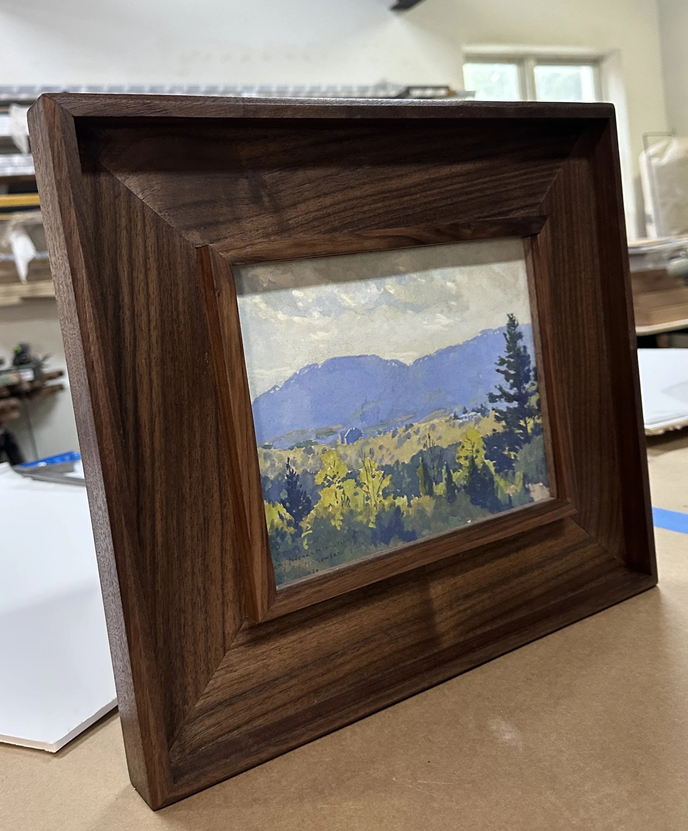 Wooden black walnut picture frame with landscape painting inside, placed on beige surface, with blurred storage shelves and window in background.