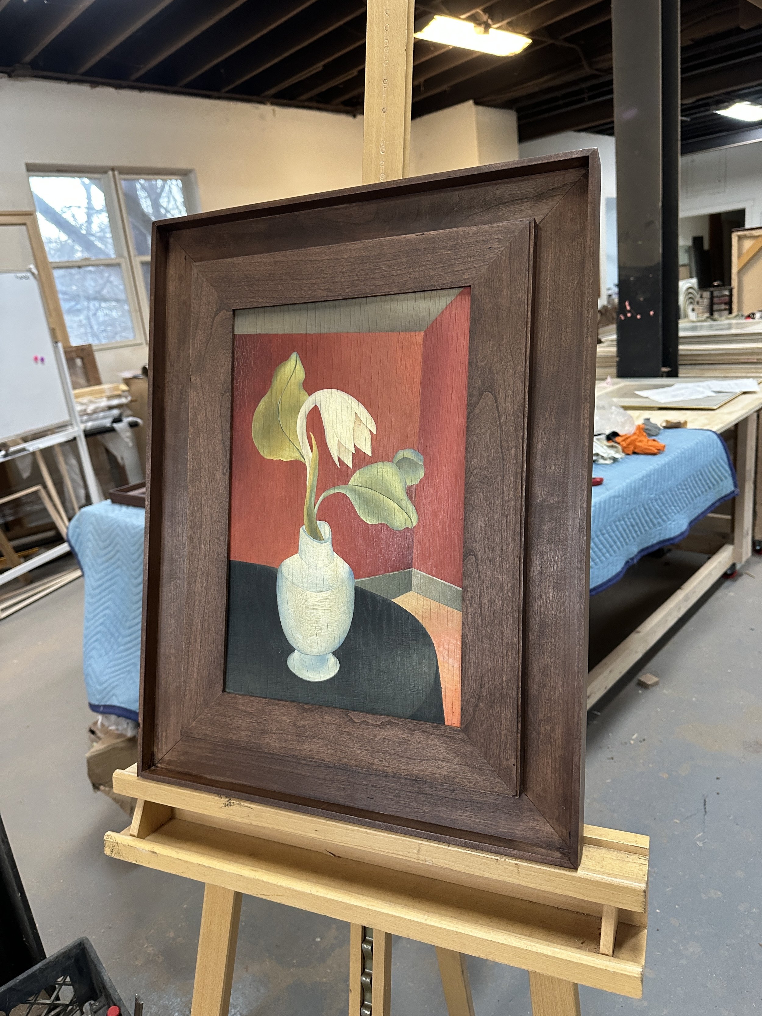 Hand crafted dark stained cherry step framed painting of a white vase with a green leafed plant inside, set against a warm background, with a dark tabletop base.