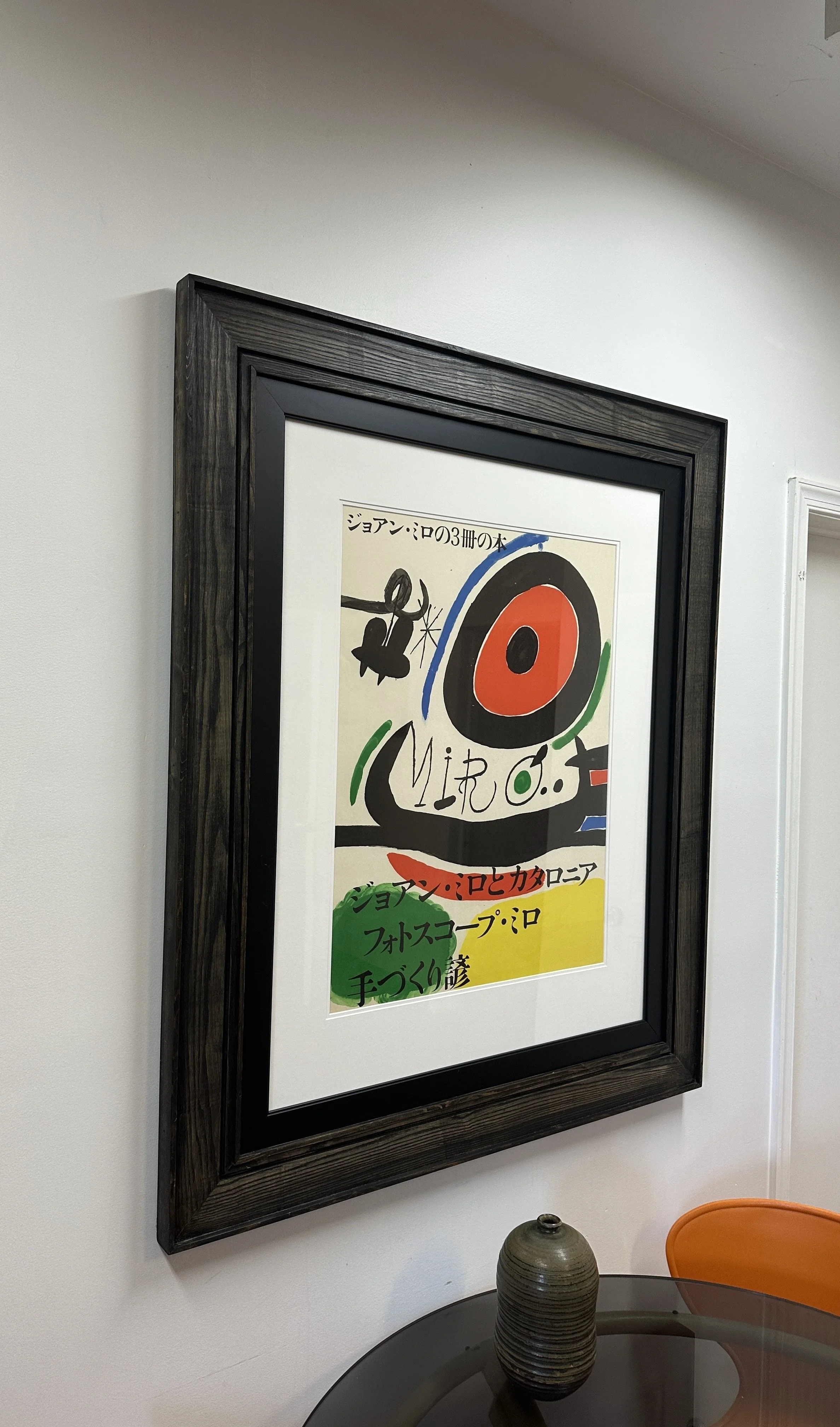 A black contemporary step framed poster with colorful abstract art hanging on a white wall, next to a black table with a decorative vase and an orange chair.