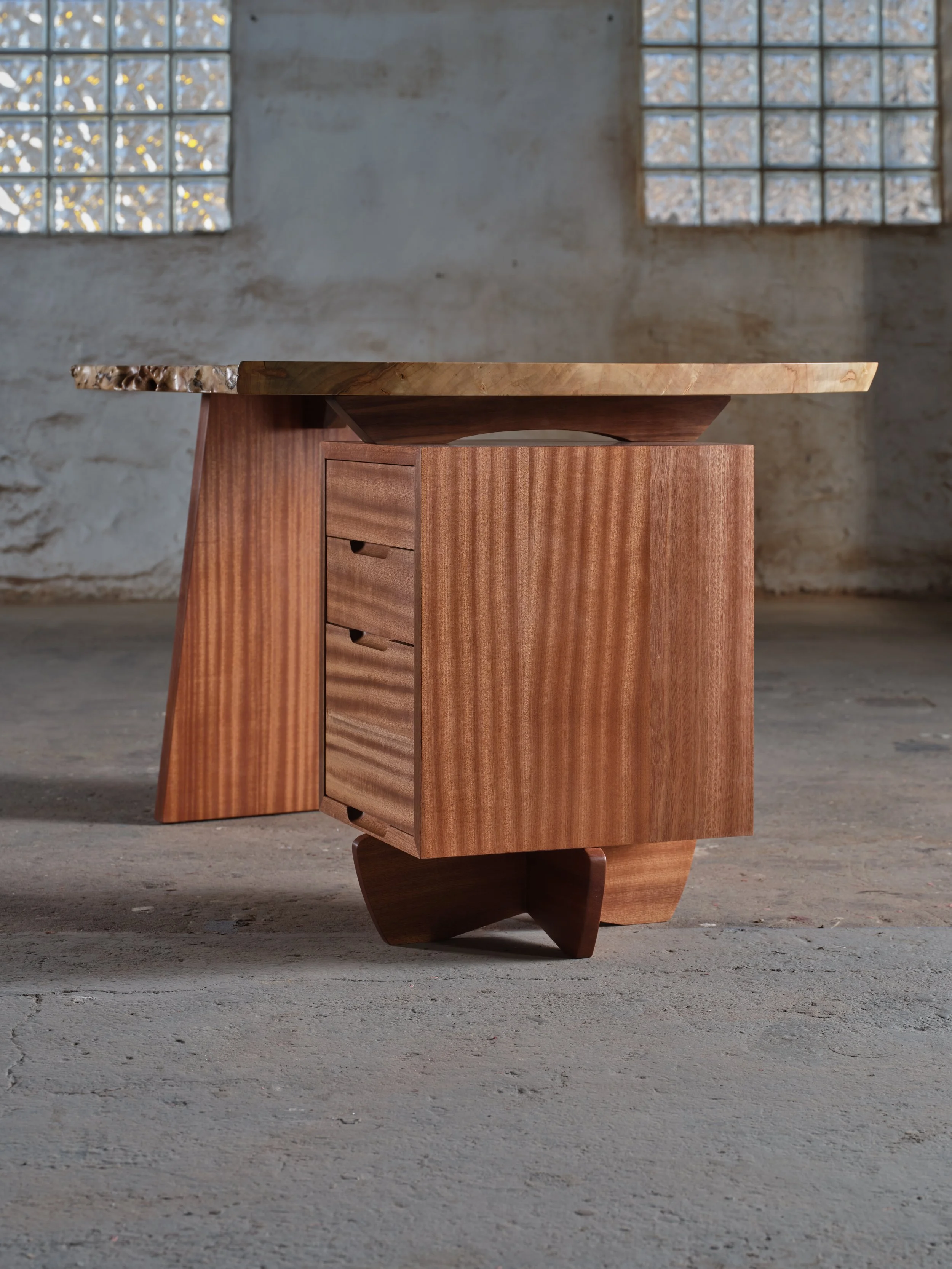 A piece of furniture made of wood with a flat top surface and a small drawer, standing on a concrete floor with a textured, unfinished wall and glass block windows in the background.