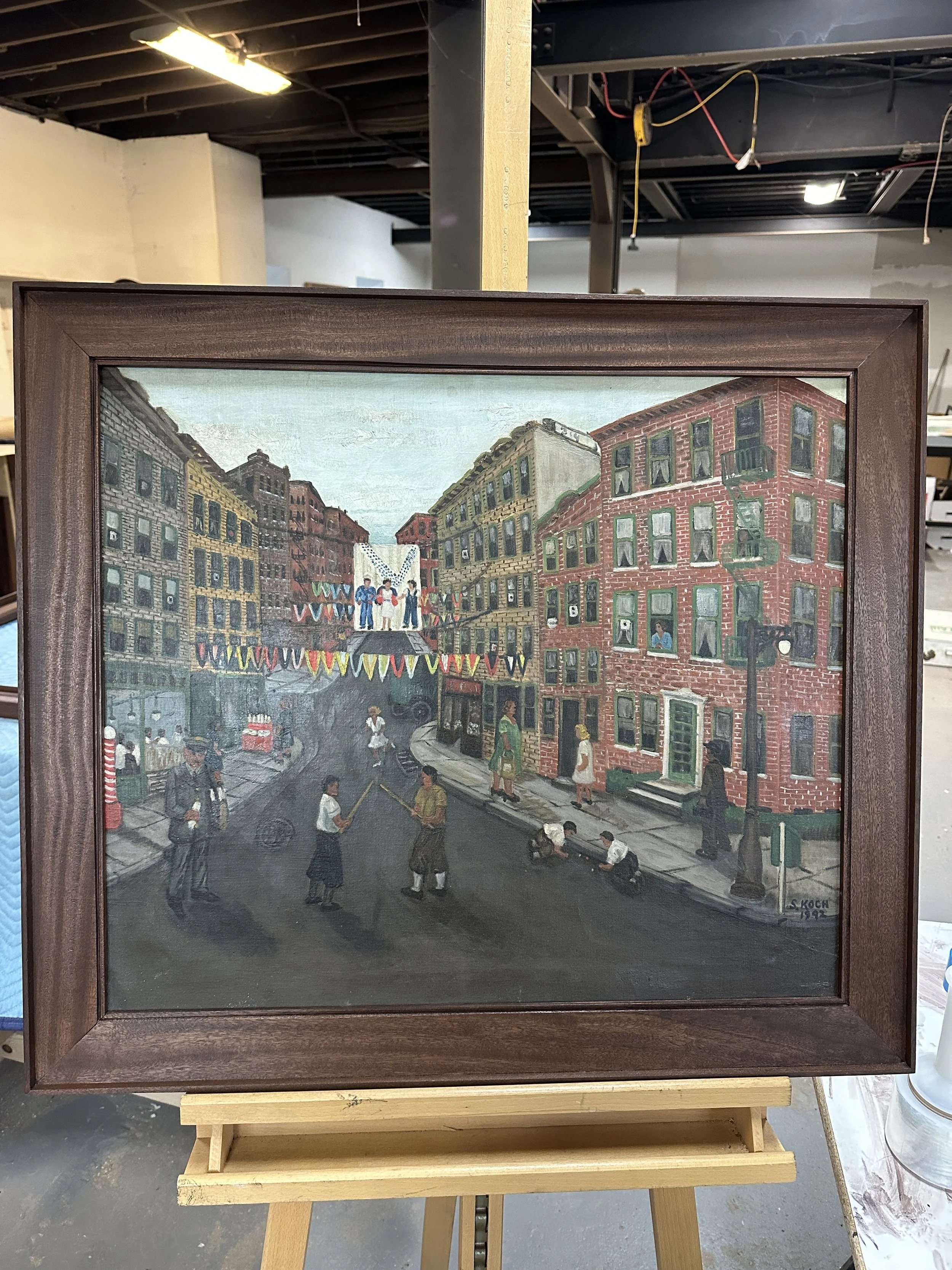 Painting of a lively city street scene in Brooklyn, New York, with colorful buildings, people walking, and festive decorations hanging above the street. Framed in hand crafted sapele step frame.