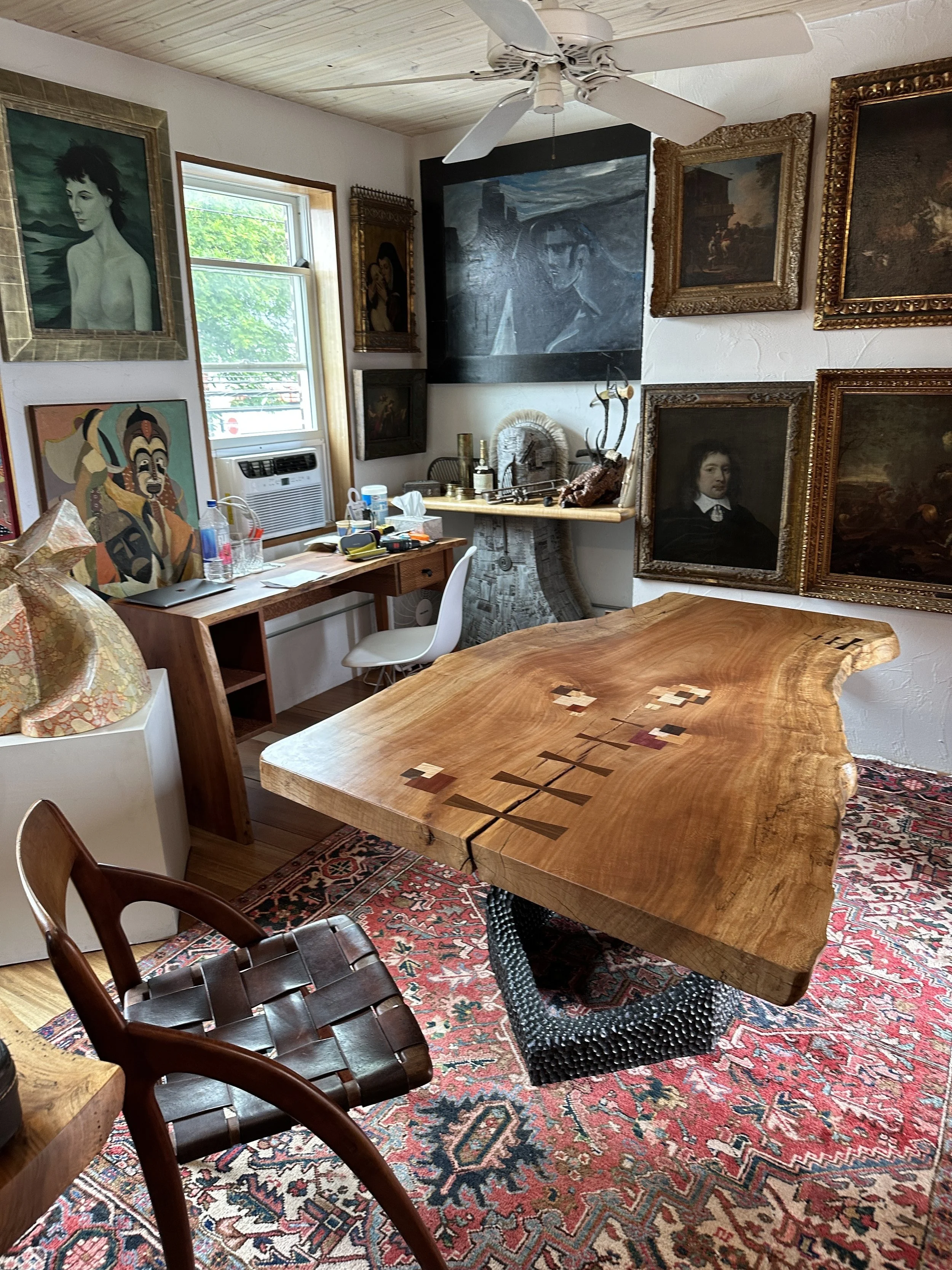 A cozy room with a large wooden table, surrounded by various chairs, and decorated with multiple framed paintings on the walls. There's a window with an air conditioning unit, a patterned rug, and a ceiling fan above.