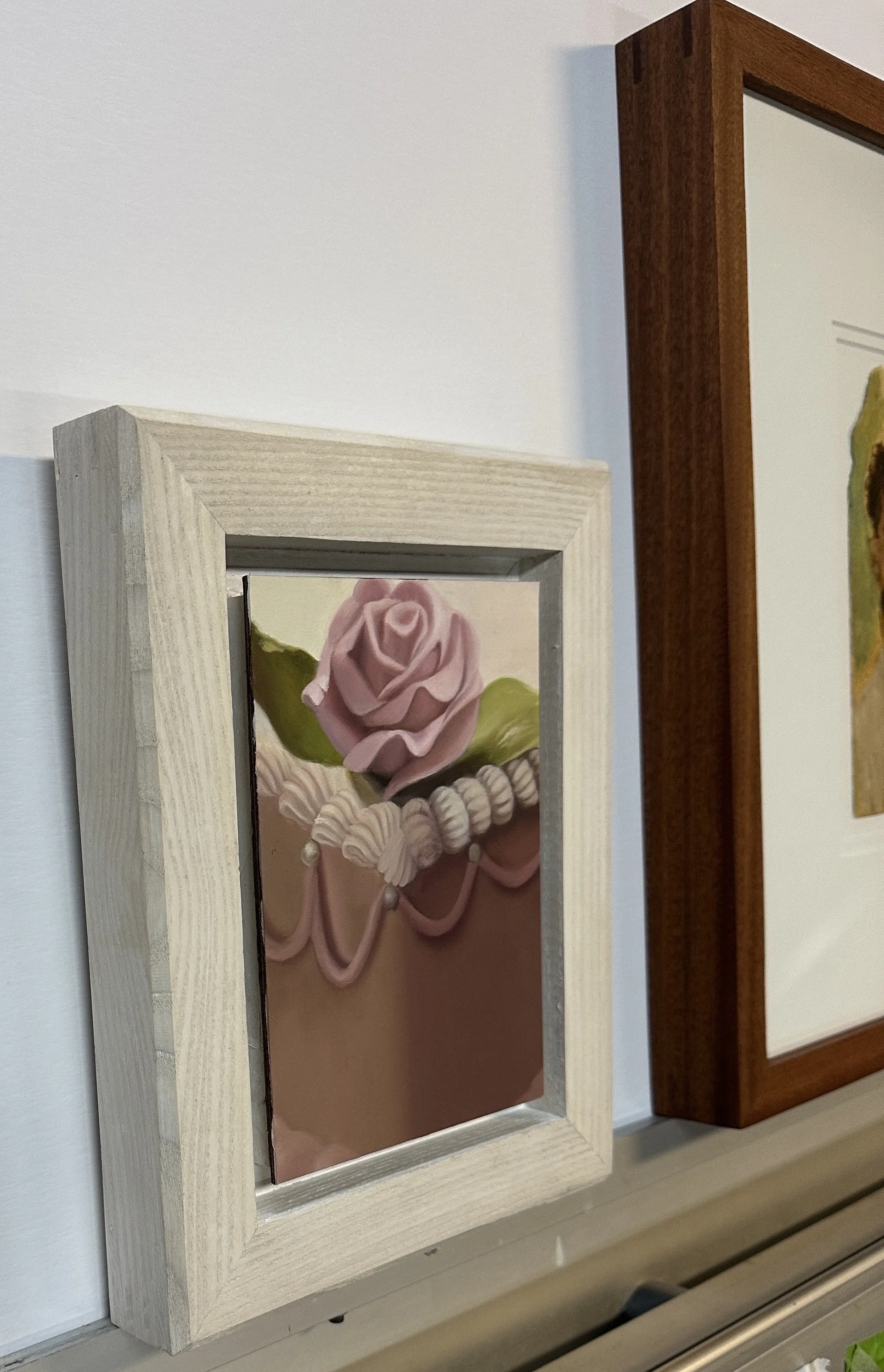 Close-up of three framed pictures hanging on a white wall, featuring colorful paintings of roses, one in a light wood frame, and two in darker wood frames.
