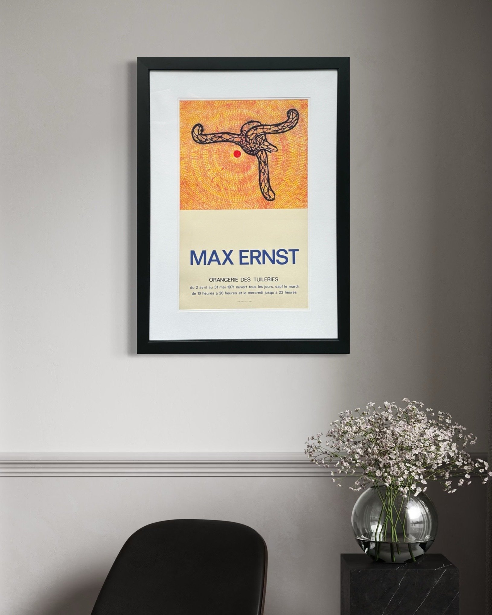 Interior scene with a framed Max Ernst art poster on the wall, a vase of white flowers on a black marble table, and a black chair in the foreground.