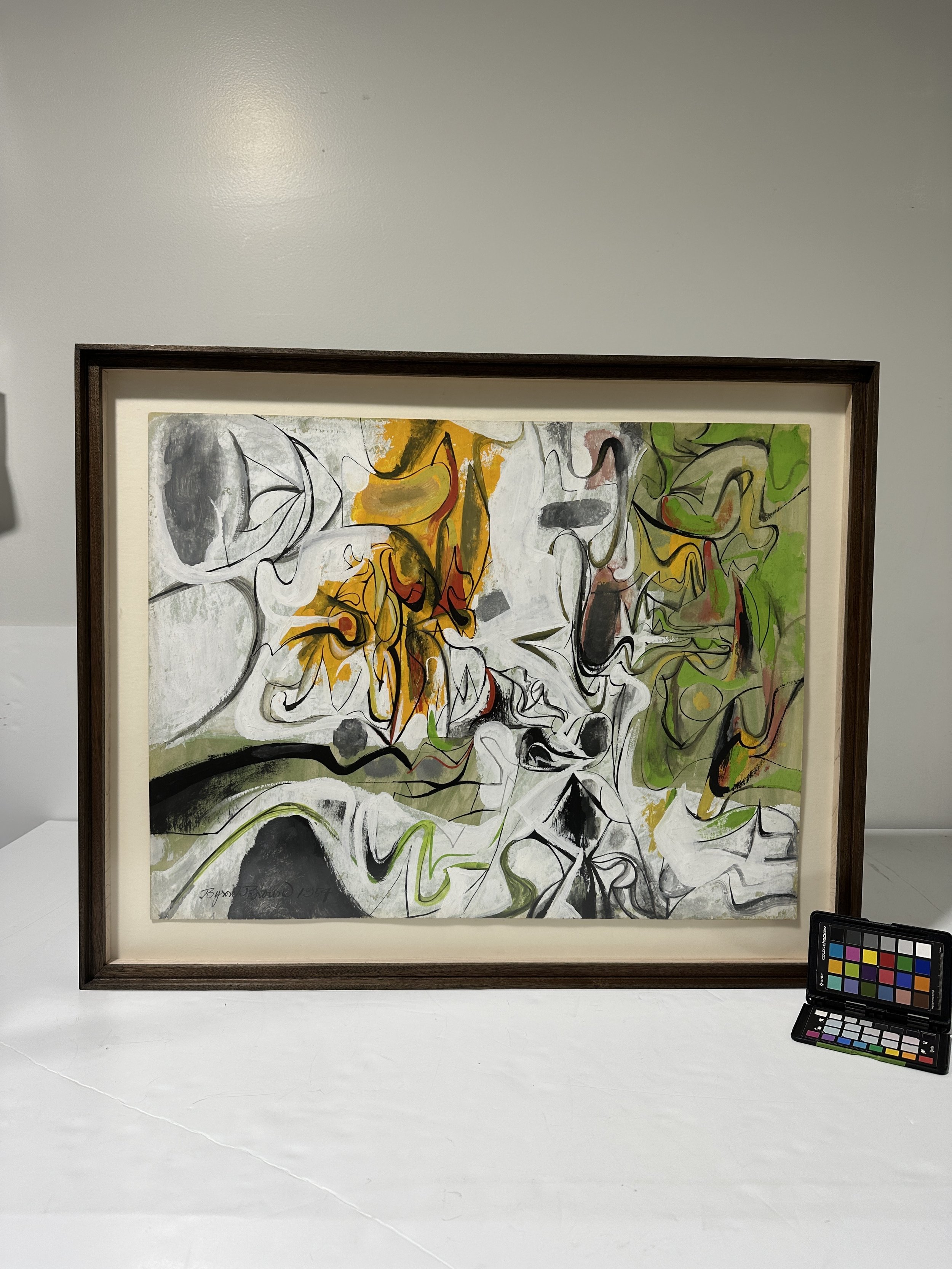 An abstract expressionist painting, framed in stained mahogony, featuring bold lines, shapes, and splashes of color including yellow, orange, green, black, gray, and white, framed and displayed on a white wall with a color calibration chart nearby.