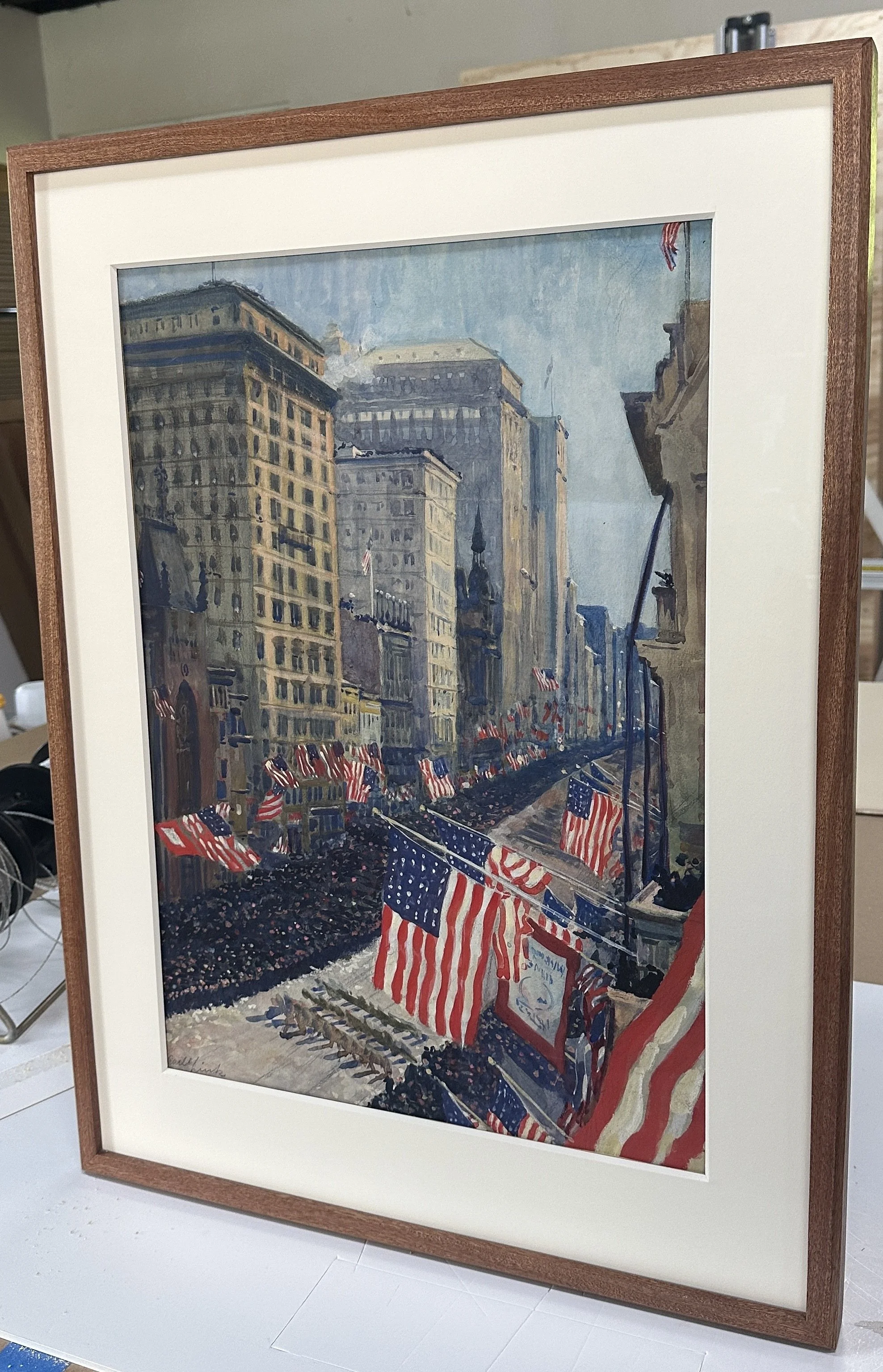 A painting of a street scene during a parade with American flags, tall buildings, and crowd banners in a cityscape in crafted mahogany frame.